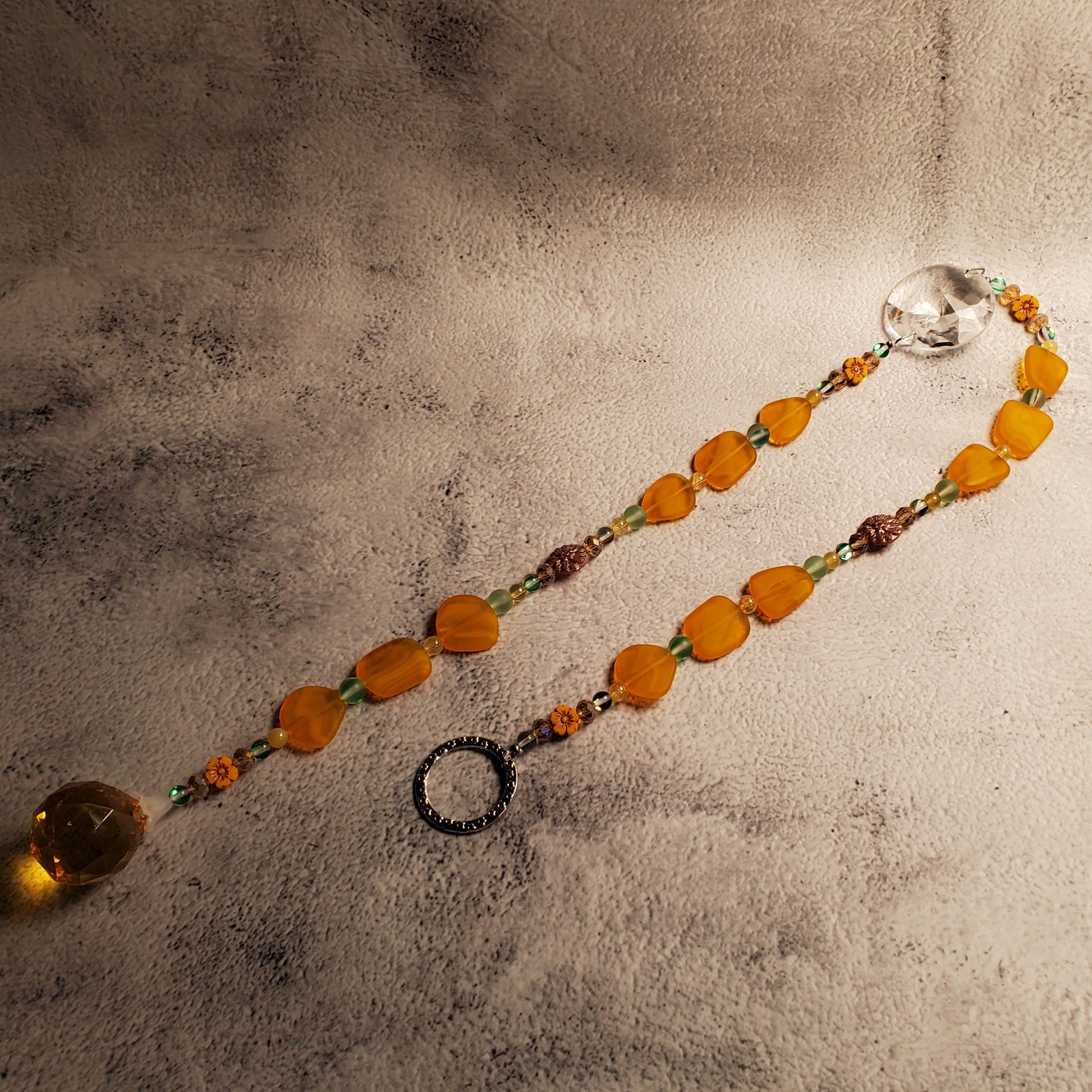 A beaded suncatcher with orange and yellow sea glass beads, green accents, and a clear faceted crystal, displayed on a textured gray surface.