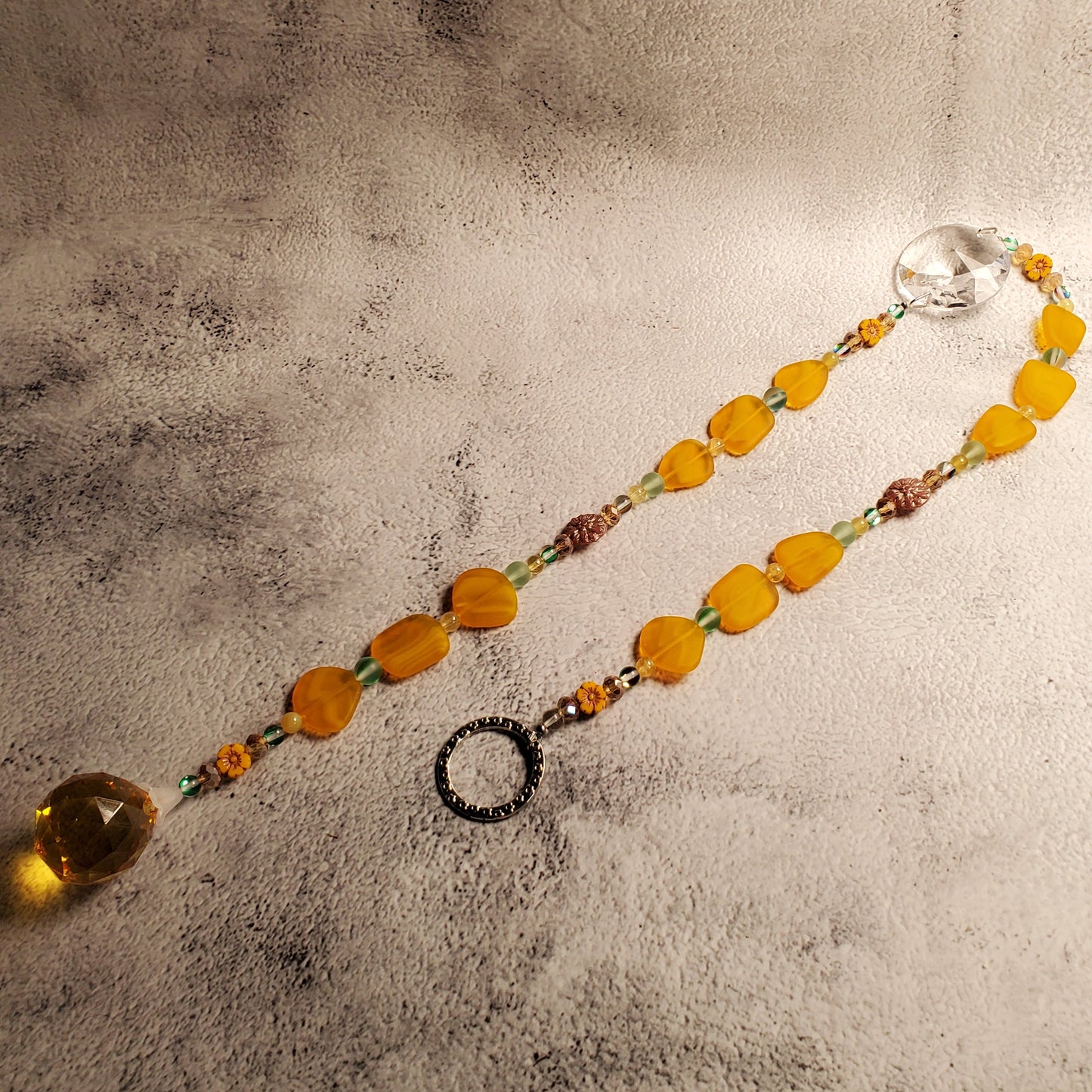 A beaded suncatcher with translucent orange-yellow sea glass beads, small yellow and green spacer beads, and a large faceted clear crystal pendant, displayed on a textured gray surface.