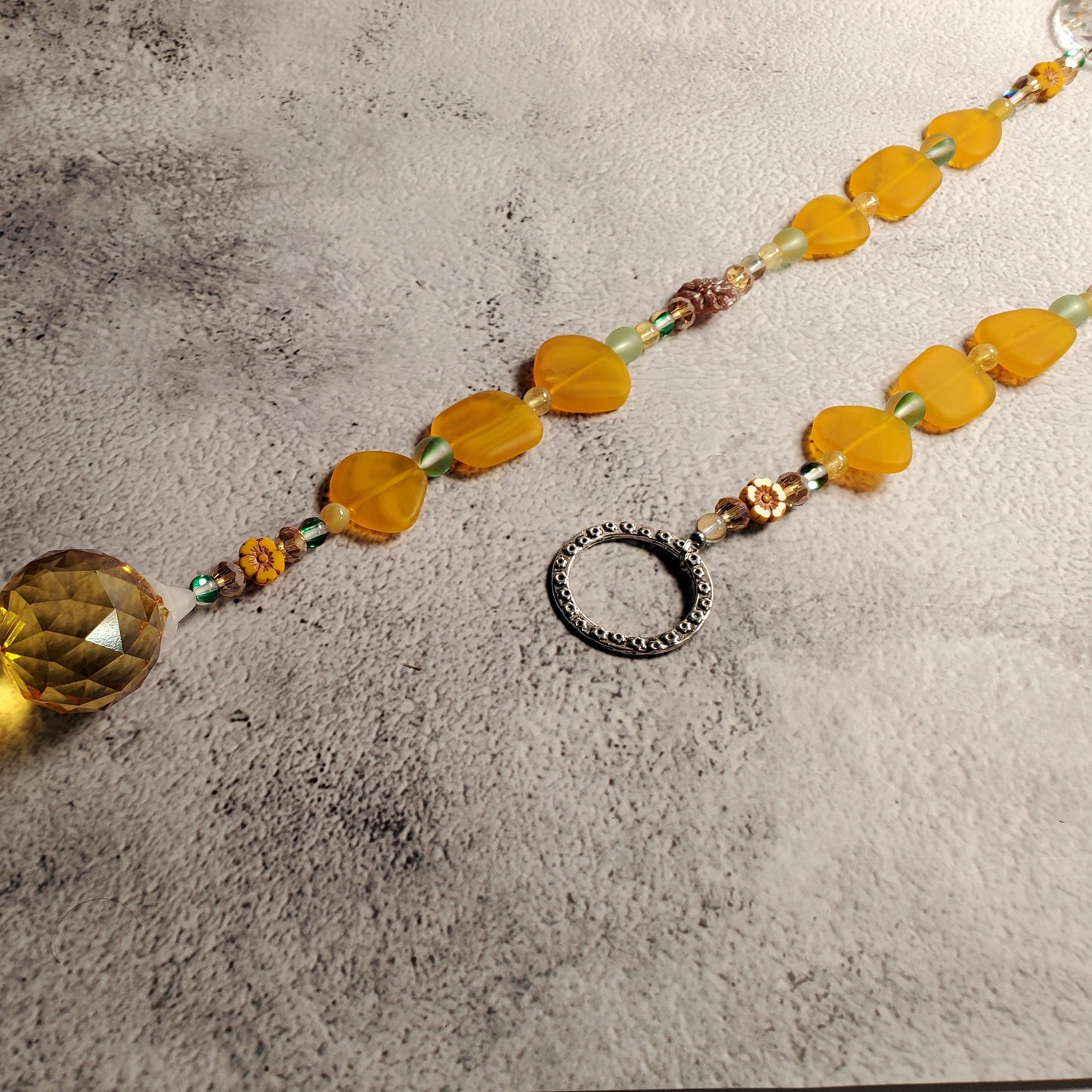 A beaded suncatcher with sea glass yellow-orange beads, a faceted yellow-amber crystal prism, and a textured silver ring, displayed on a gray textured surface.
