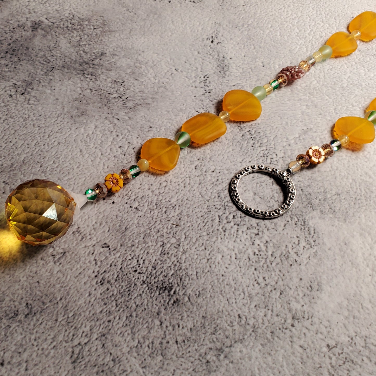 A beaded suncatcher with translucent orange-yellow sea glass beads, a faceted round amber-yellow prism, and small multicolor spacer beads, laid on a textured gray surface next to a silver ring hanger.
