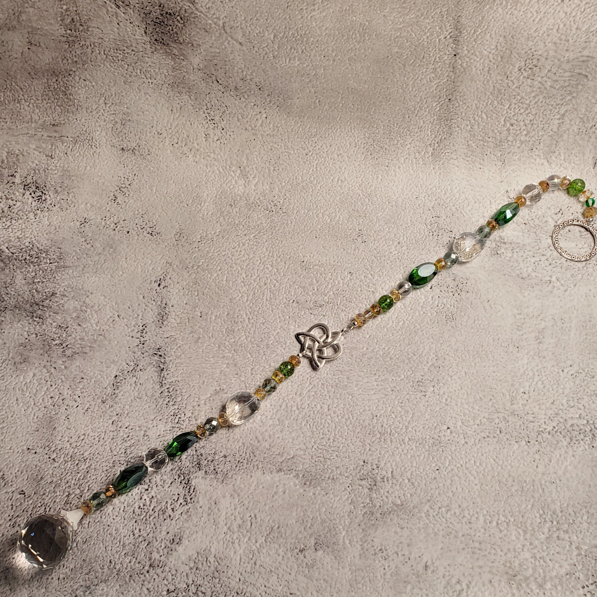 A beaded suncatcher with green and clear faceted beads, a silver Celtic knot heart centerpiece, and a large clear crystal pendant, laid diagonally on a textured gray surface.