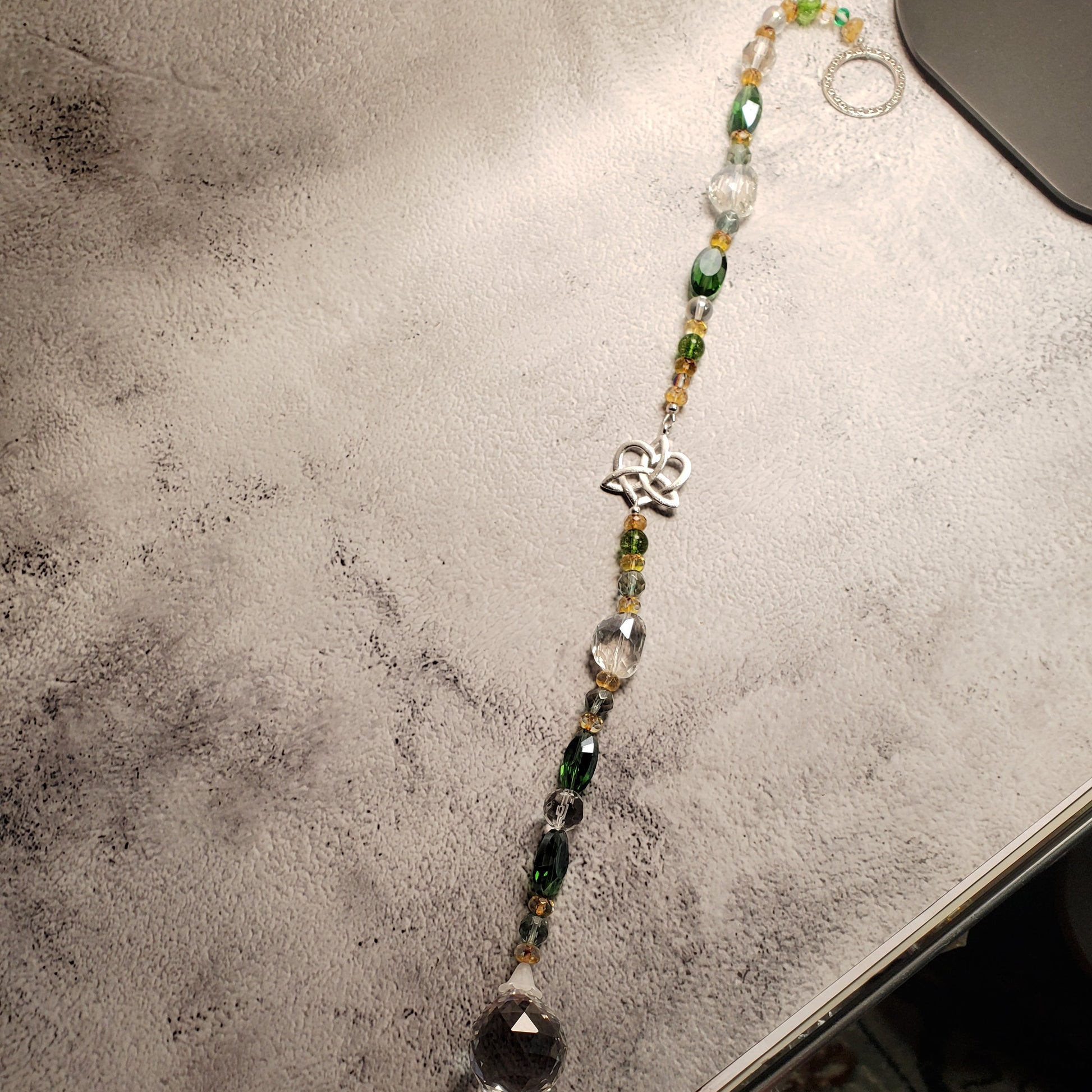 A beaded suncatcher with green, yellow, and clear faceted beads, a silver Celtic knot heart charm, and a large clear teardrop crystal pendant, displayed on a textured gray surface.