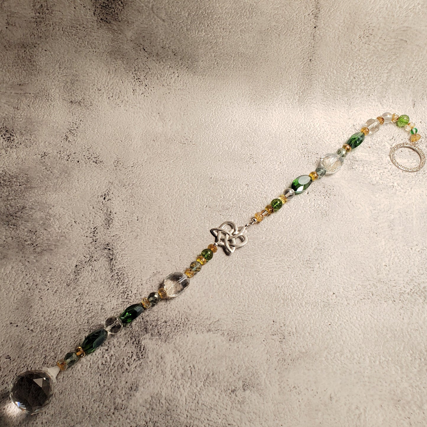 A beaded suncatcher with green, clear, and gold-toned beads, a silver-toned Celtic knot heart charm, and a large clear faceted bead at the end, displayed on a textured gray backdrop.