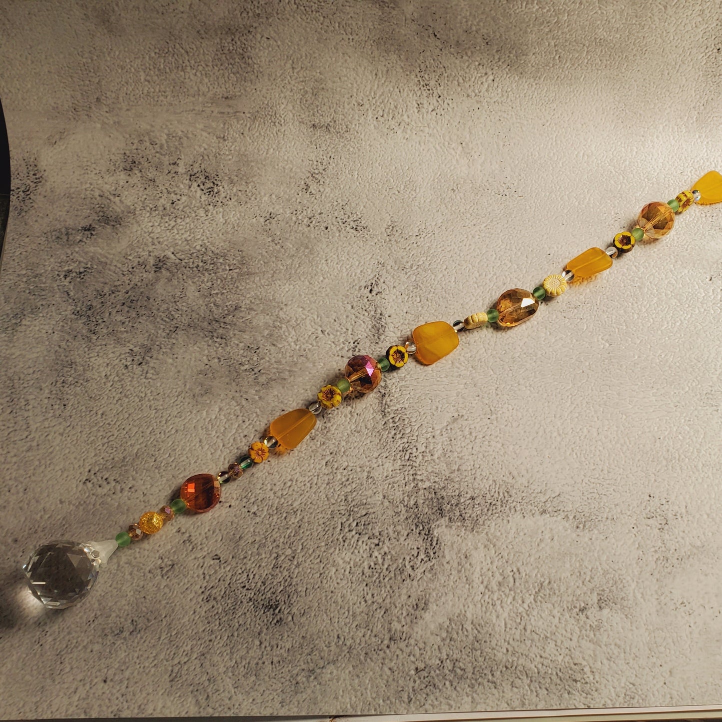 A beaded suncatcher with amber, orange, and yellow faceted beads and a clear crystal ball at one end, laid diagonally on a textured light gray backdrop.