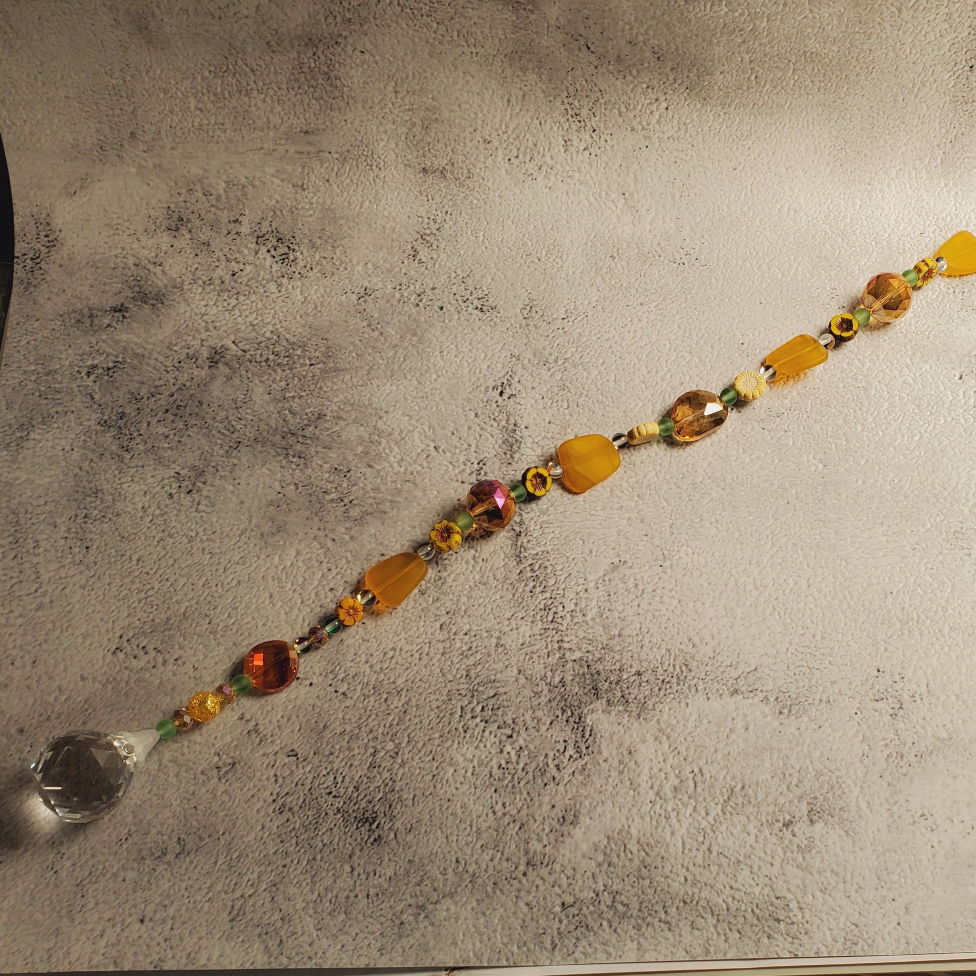 A beaded suncatcher with amber, orange, and yellow faceted beads and a clear crystal ball at one end, laid diagonally on a textured light gray backdrop.