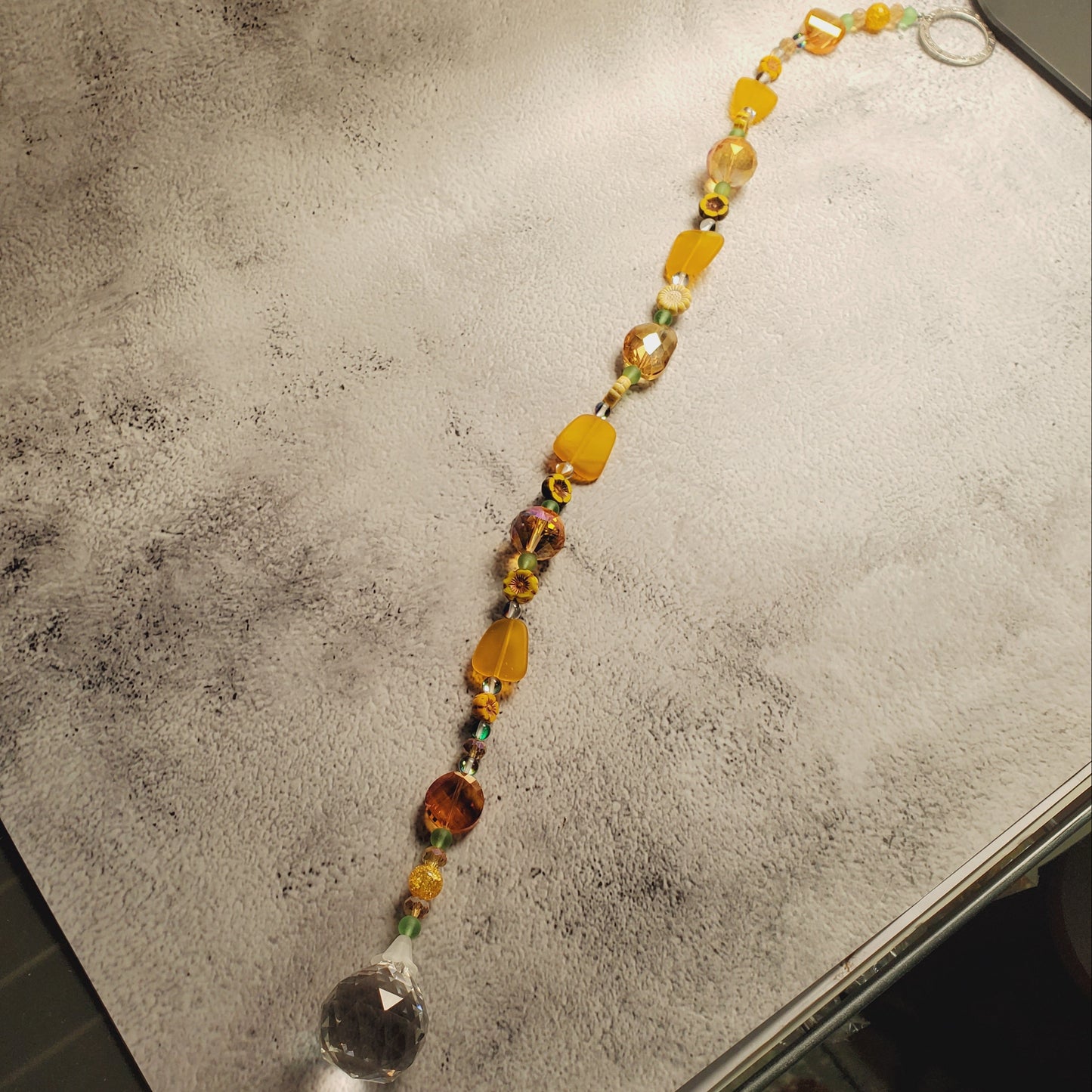 A beaded suncatcher with yellow, orange, and clear faceted beads, laid diagonally on a textured white surface.