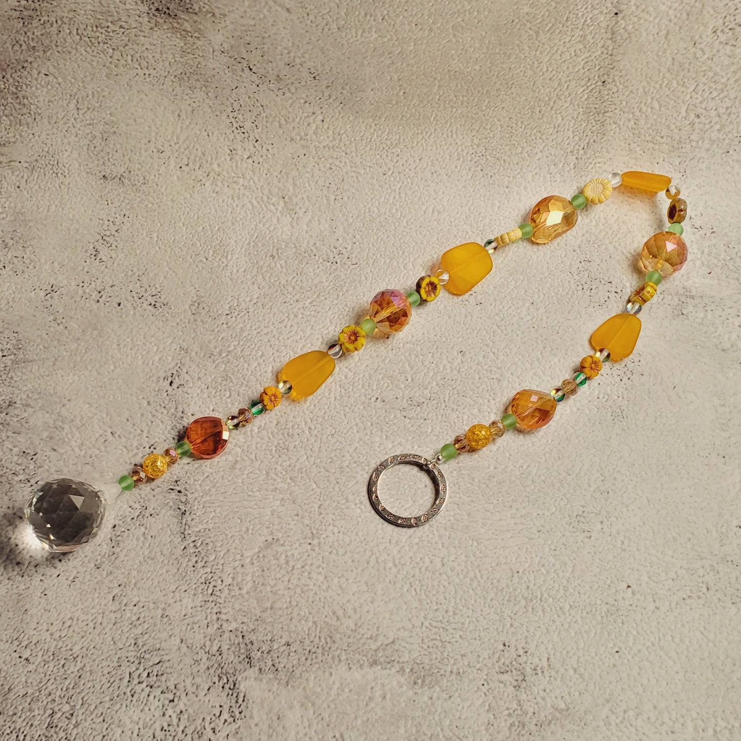 A beaded suncatcher with sea glass yellow and orange beads, small green accents, a silver metal ring, and a clear crystal pendant, laid on a textured gray surface.