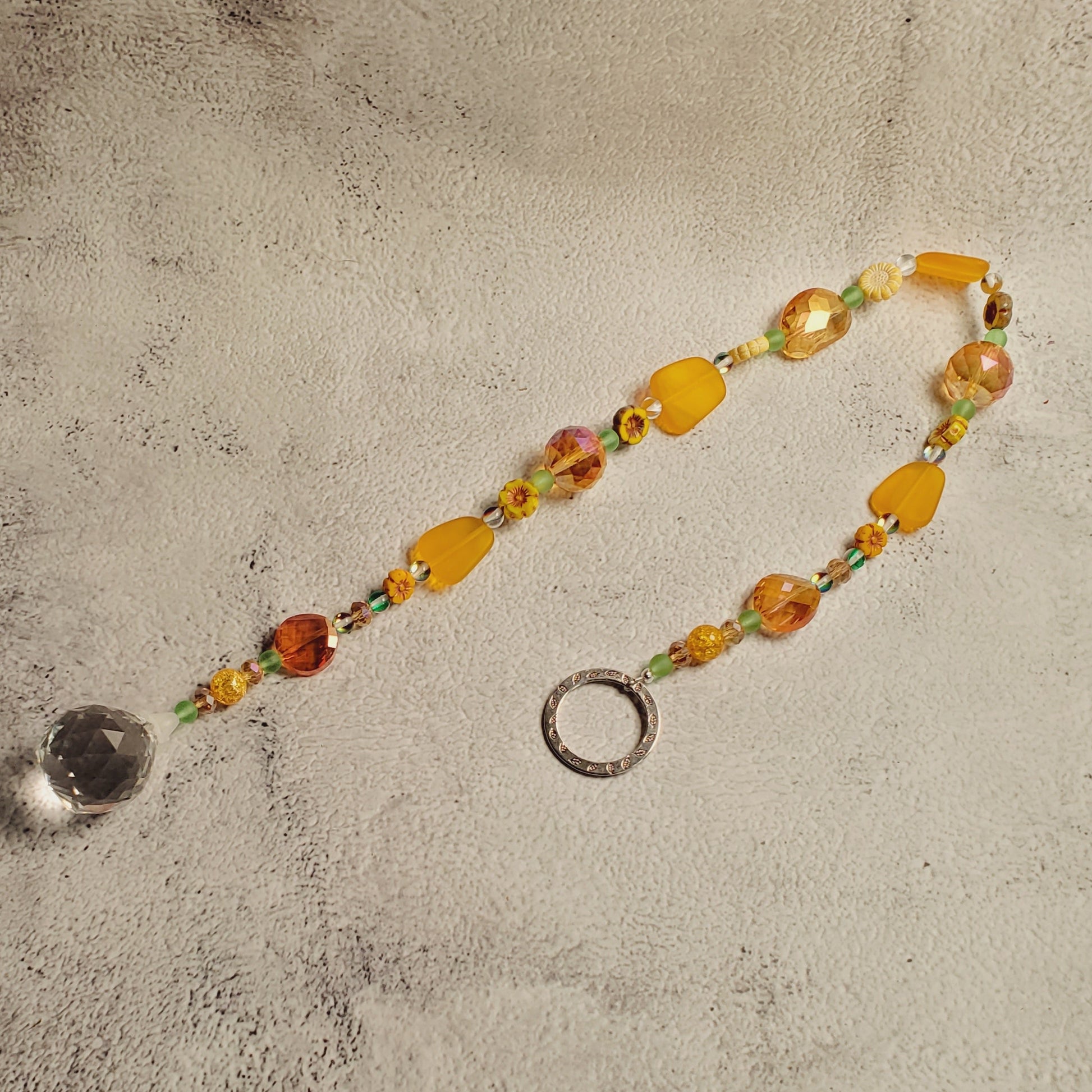 A beaded suncatcher with sea glass yellow and orange beads, small green accents, a silver metal ring, and a clear crystal pendant, laid on a textured gray surface.