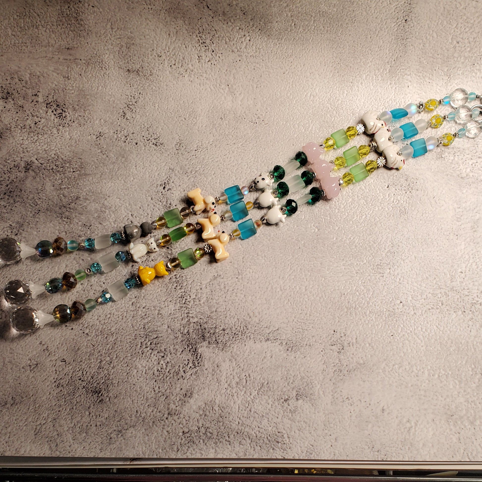 Handmade beaded suncatchers laid out diagonally on a textured gray tabletop. The piece features a mix of colorful glass beads in turquoise, green, yellow, white, and brown, alongside clear crystal prisms.