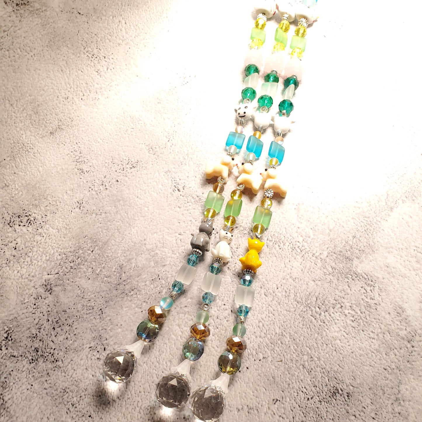 A beaded decorative suncatcher with translucent and opaque glass beads in green, blue, yellow, white, and brown, arranged diagonally on a textured white surface.