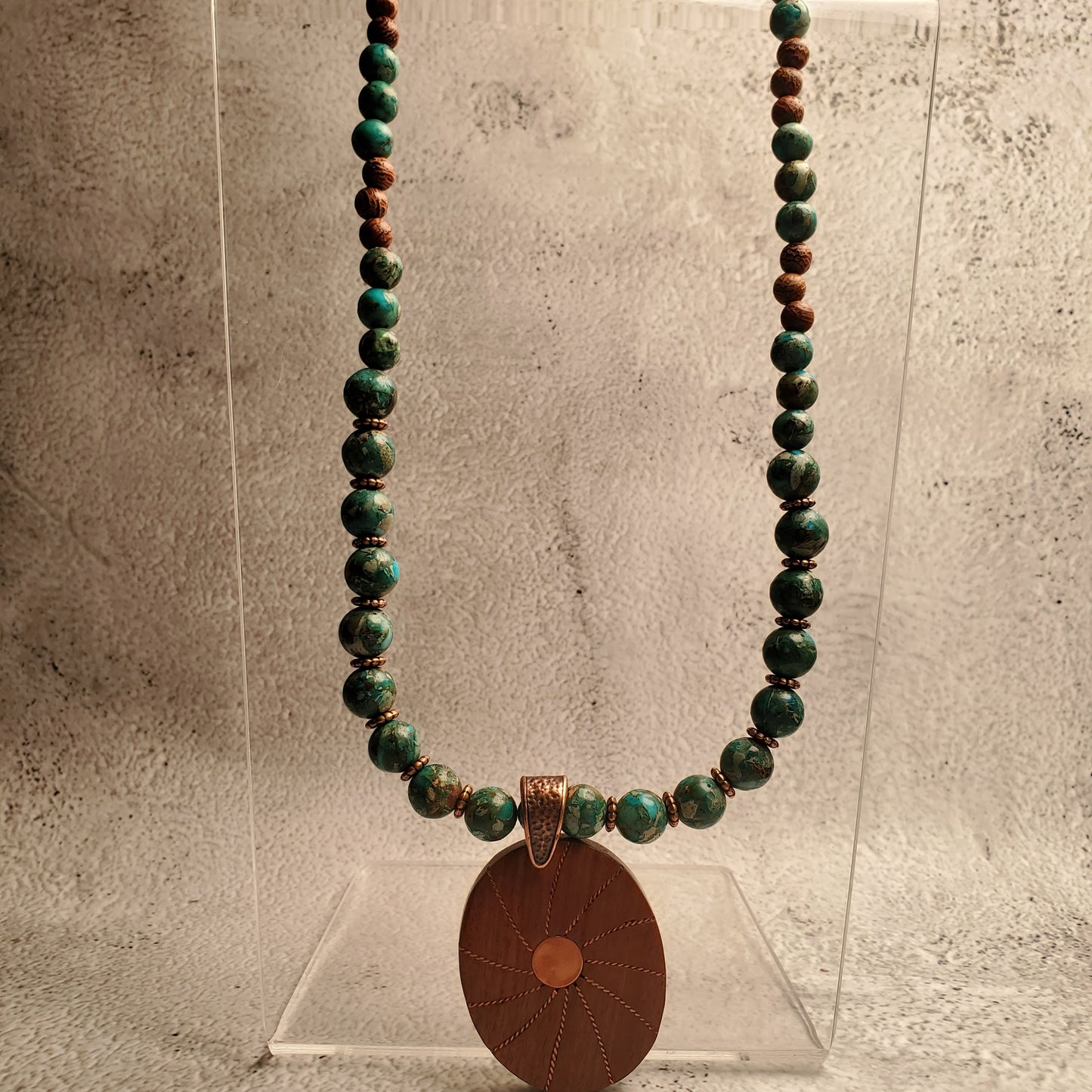 A beaded necklace with green and brown beads and a large oval brown pendant, displayed on a clear acrylic stand against a textured gray background.