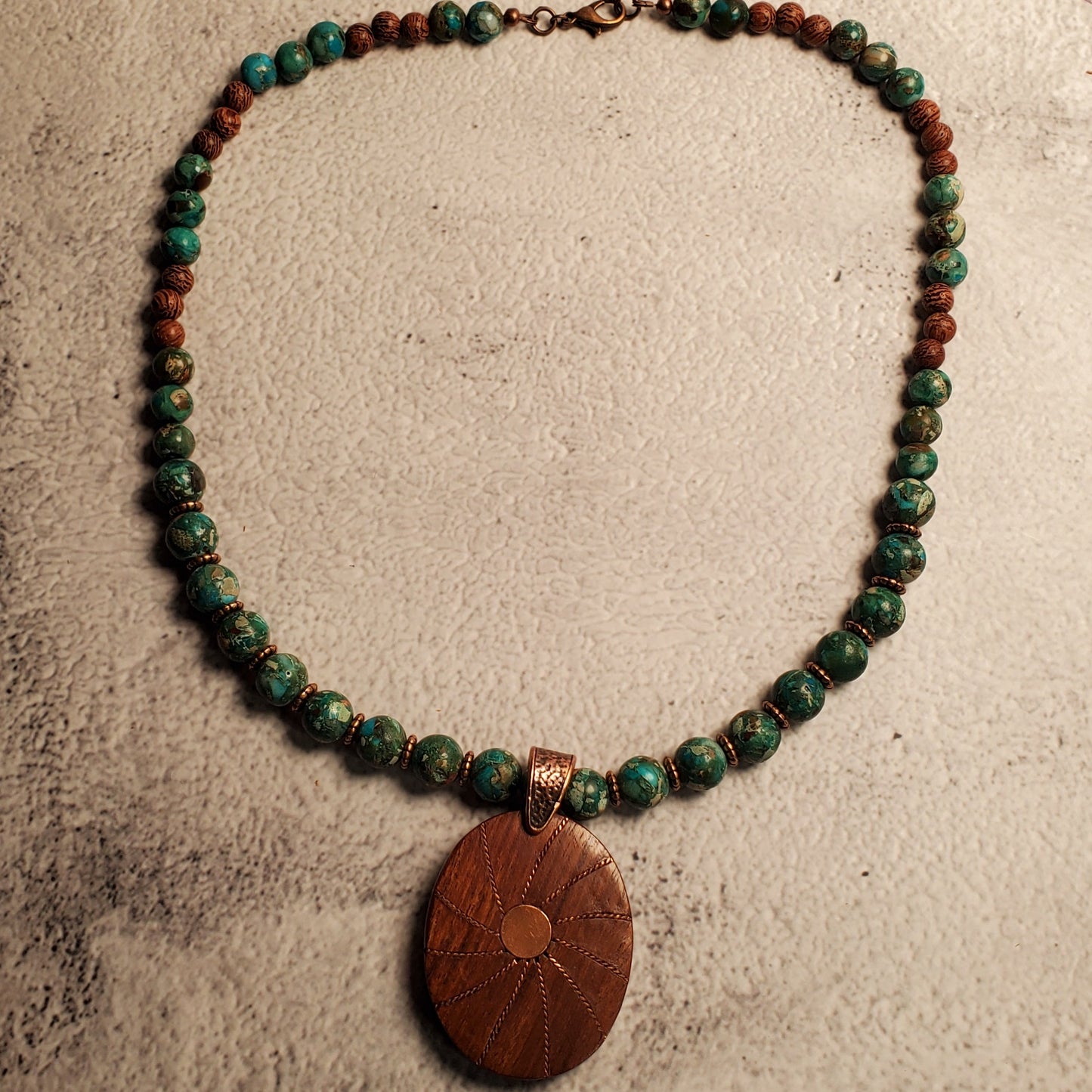 A beaded necklace with green and reddish-brown beads and a large oval brown wooden pendant, displayed on a textured beige surface.