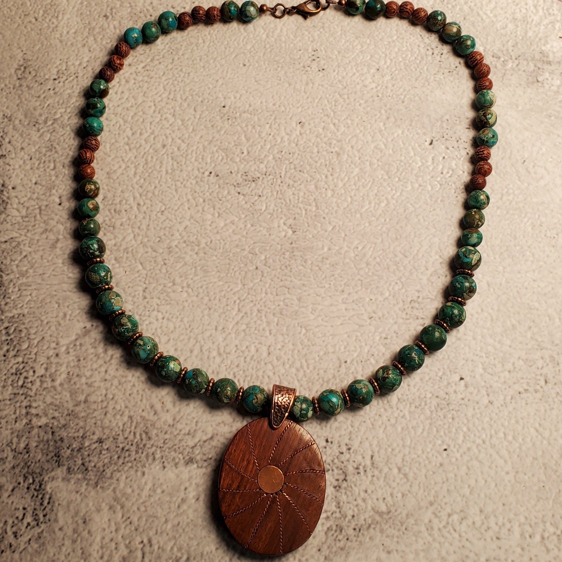 A beaded necklace with green and reddish-brown beads and a large oval brown wooden pendant, displayed on a textured beige surface.