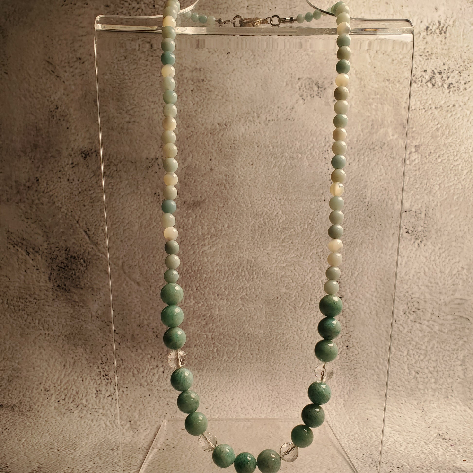 A beaded necklace made of green and white round beads, displayed on a clear acrylic stand against a textured gray background.