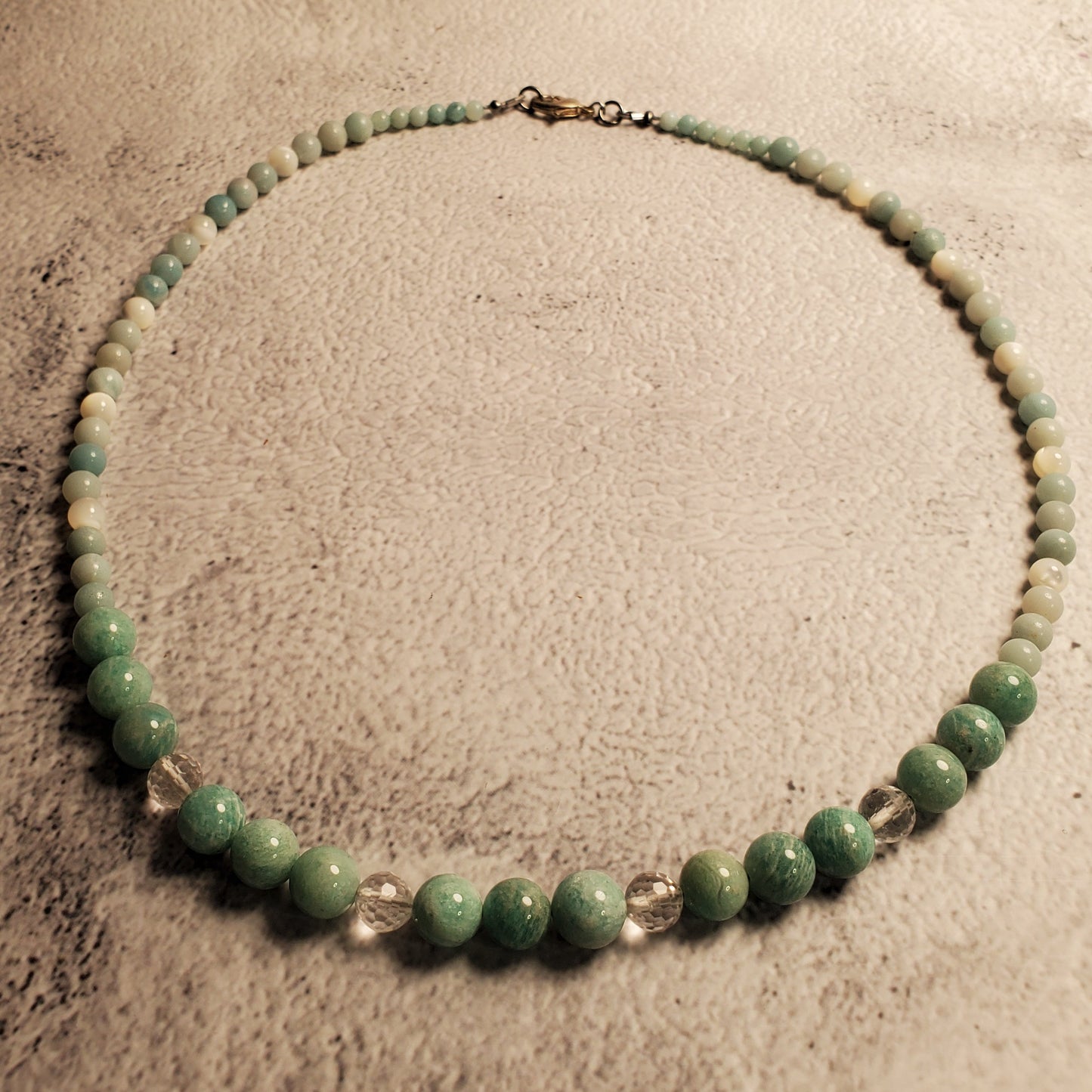 A beaded necklace made of green and light blue-green round beads with a few clear faceted accent beads, arranged in a circle on a textured beige surface.