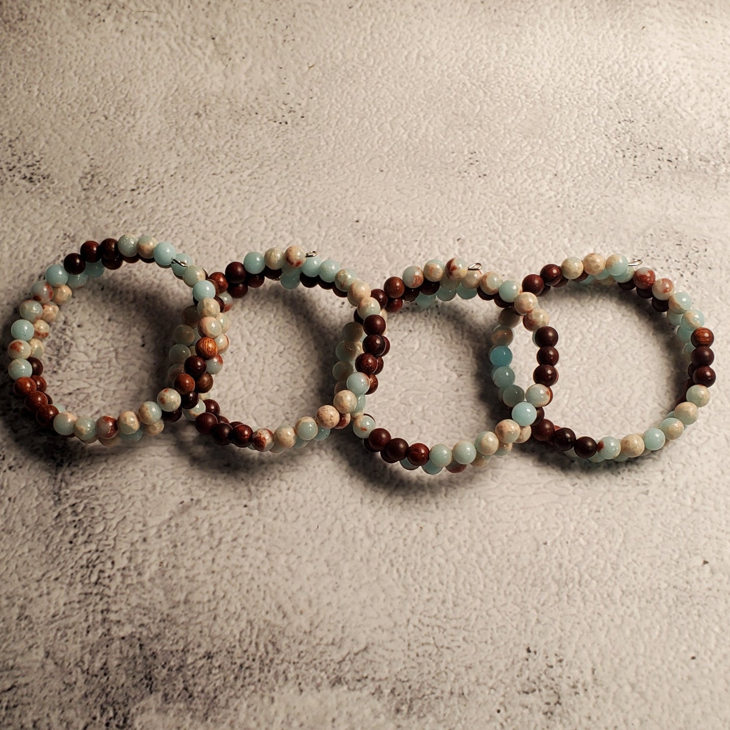 Four beaded memory wire bracelets arranged in a row on a textured gray background. The bracelets feature alternating beads of dark brown, light blue, and cream.