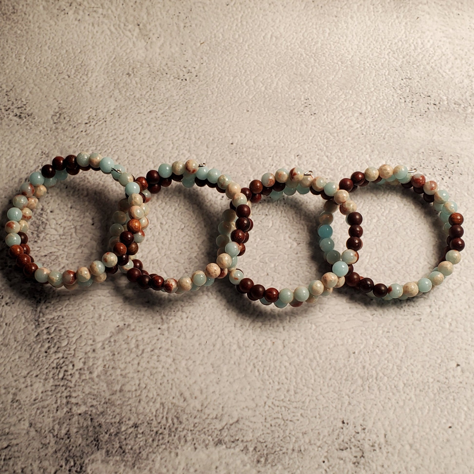Four beaded memory wire bracelets arranged in a row on a textured gray background. The bracelets feature alternating beads of dark brown, light blue, and cream.