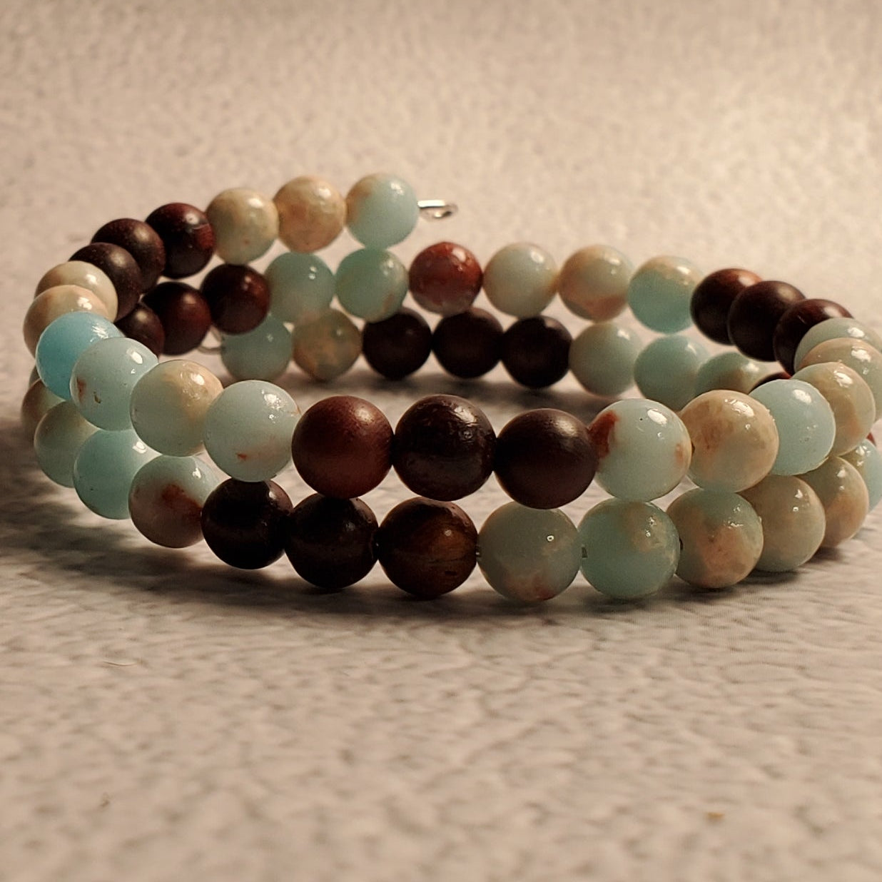 A beaded wrap bracelet with alternating light blue, dark brown, and cream beads, resting on a textured beige surface.
