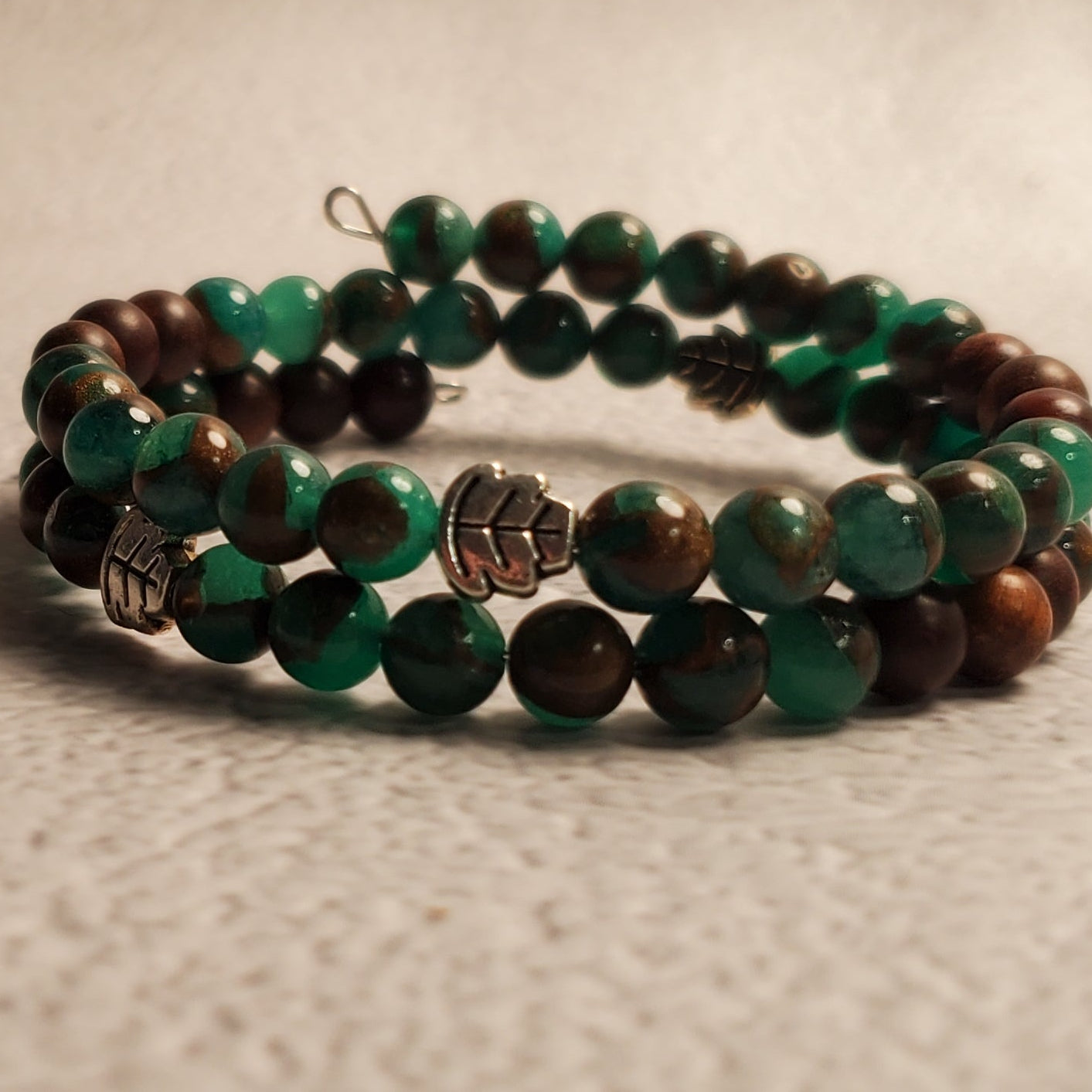 A beaded wrap bracelet featuring green and dark brown beads with a small silver-toned charm, resting on a textured beige surface.