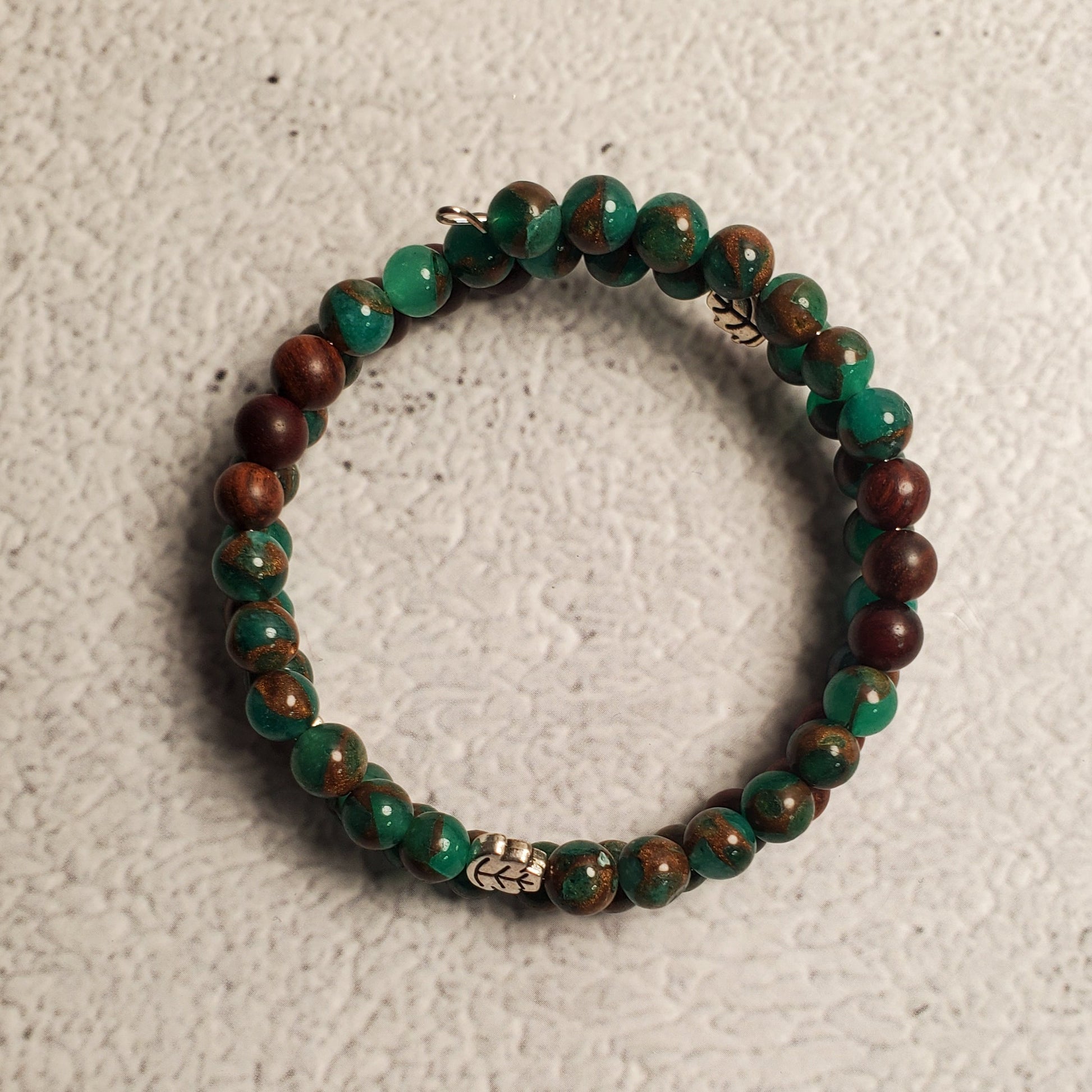 A beaded memory wire bracelet made of green and brown marbled beads with small silver spacer beads, displayed on a textured off-white surface.
