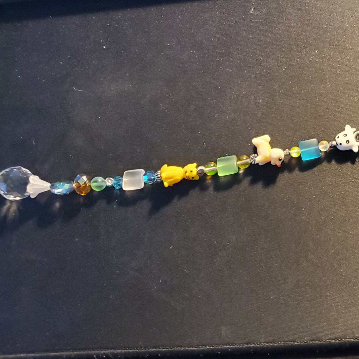 A long beaded Suncatcher laid flat on a dark desk surface, featuring multicolored translucent and opaque beads with small animal-shaped charms, plus a clear teardrop-shaped bead at the bottom. 