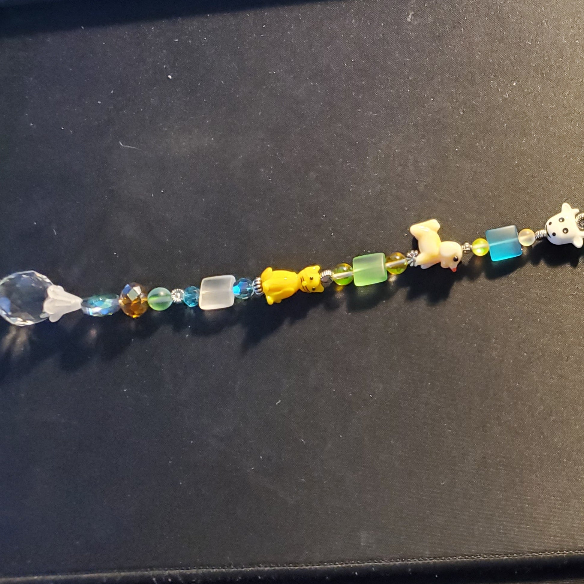 A long beaded Suncatcher laid flat on a dark desk surface, featuring multicolored translucent and opaque beads with small animal-shaped charms, plus a clear teardrop-shaped bead at the bottom. 