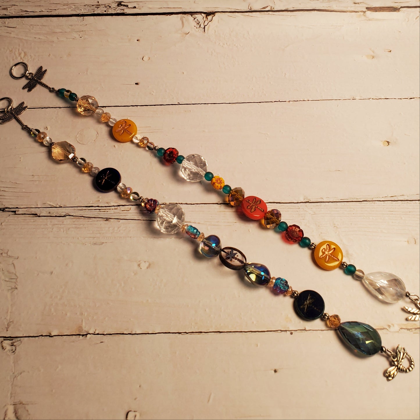 A beaded suncatcher with colorful glass beads, clear faceted beads, yellow and orange medallions, and a silver-tone metal crucifix, laid out diagonally on a rustic white-painted wooden surface.