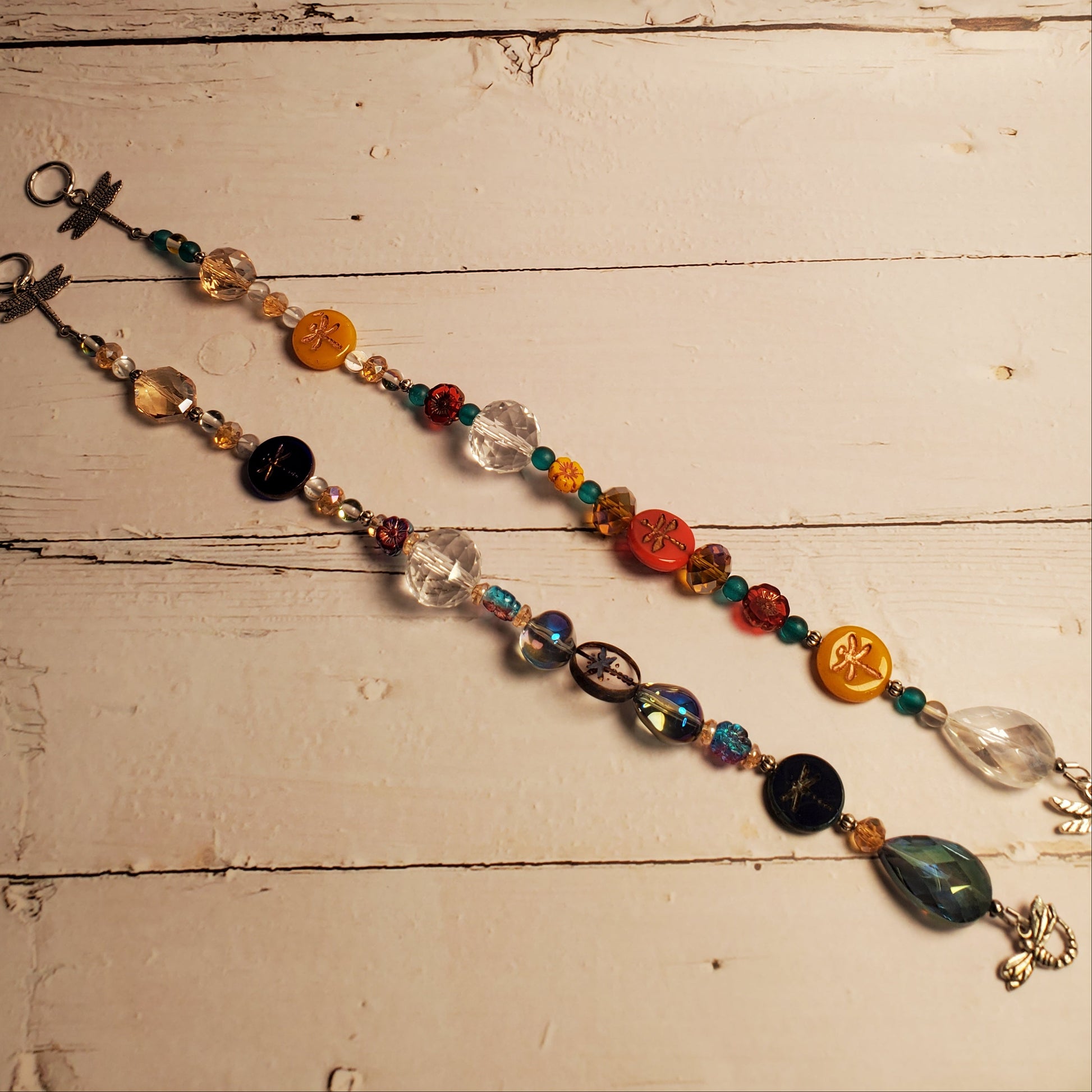 A beaded suncatcher with colorful glass beads, clear faceted beads, yellow and orange medallions, and a silver-tone metal crucifix, laid out diagonally on a rustic white-painted wooden surface.