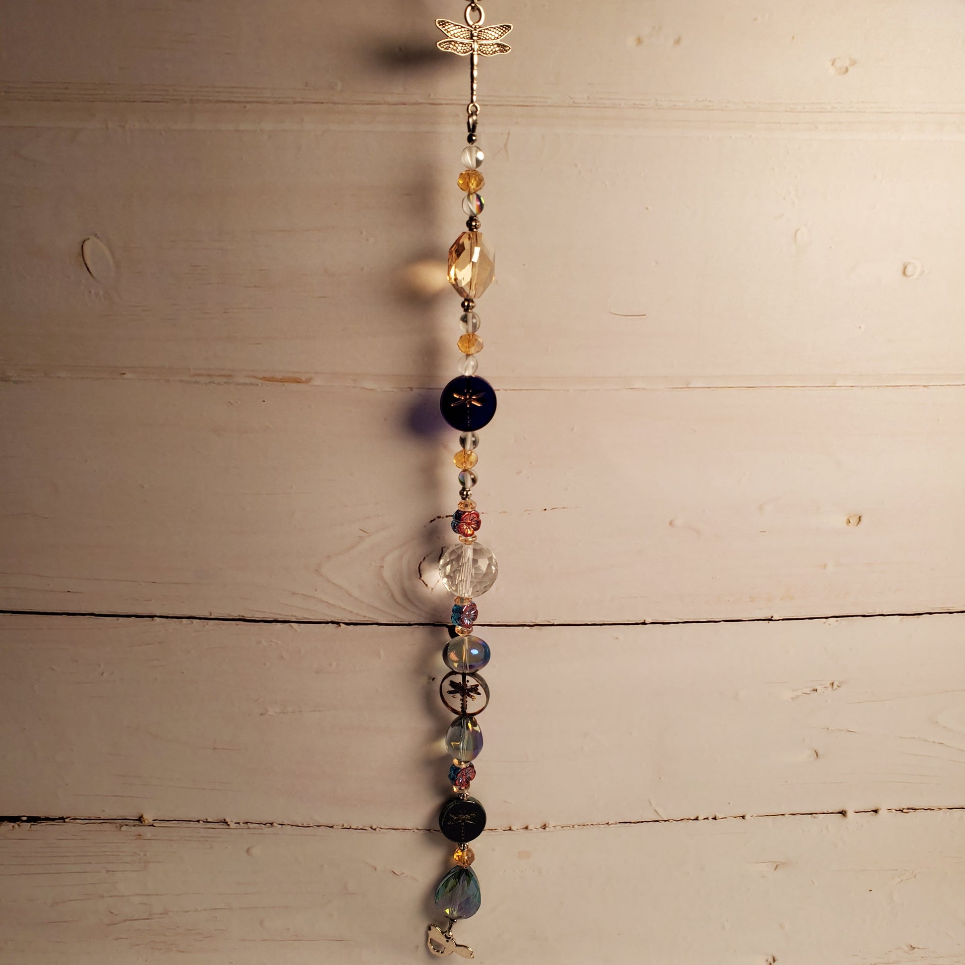 A beaded hanging suncatcher  with a small dragonfly charm at the top, suspended against a rustic white-painted wooden plank background. The hanging piece features multiple beads in black, clear, amber, blue, and metallic gold tones.