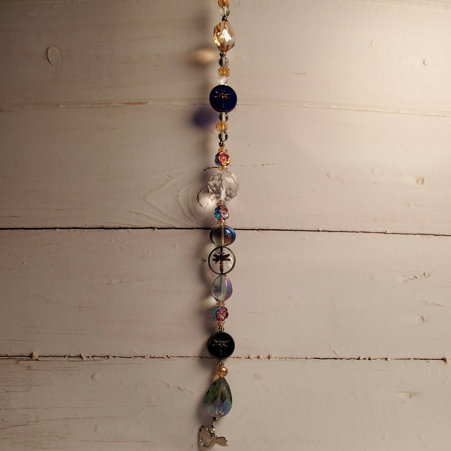 A beaded hanging suncatcher with assorted glass beads in blue, black, clear, and iridescent colors, hanging against a rustic white-painted wooden wall.