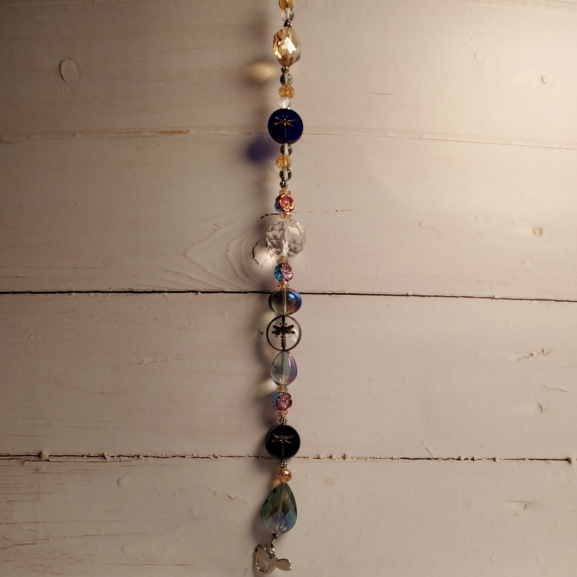 A beaded hanging suncatcher with assorted glass beads in blue, black, clear, and iridescent colors, hanging against a rustic white-painted wooden wall.