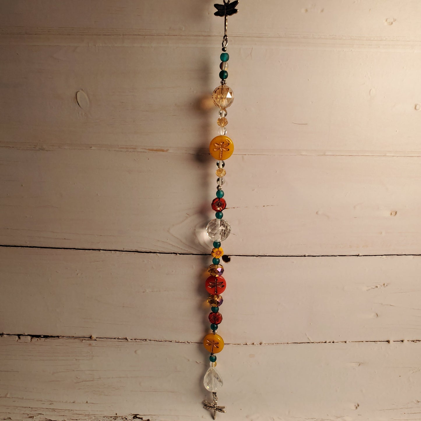A beaded suncatcher with colorful glass beads, a black dragonfly charm, and clear glass elements, hanging against a rustic white wooden plank wall.