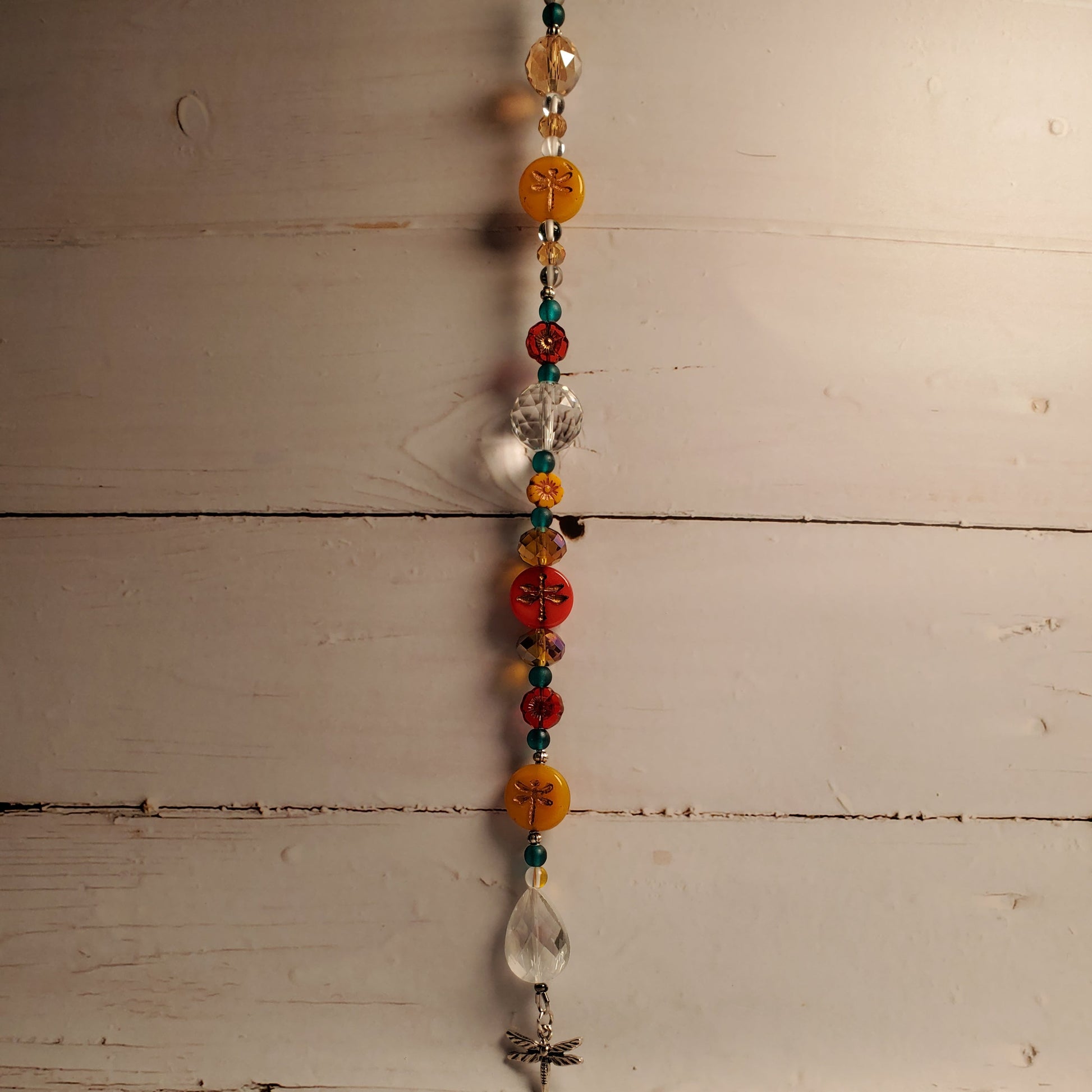 A beaded hanging suncatcher with colorful round and faceted beads, including orange, red, teal, and clear glass, suspended against a rustic white-painted wooden plank background.