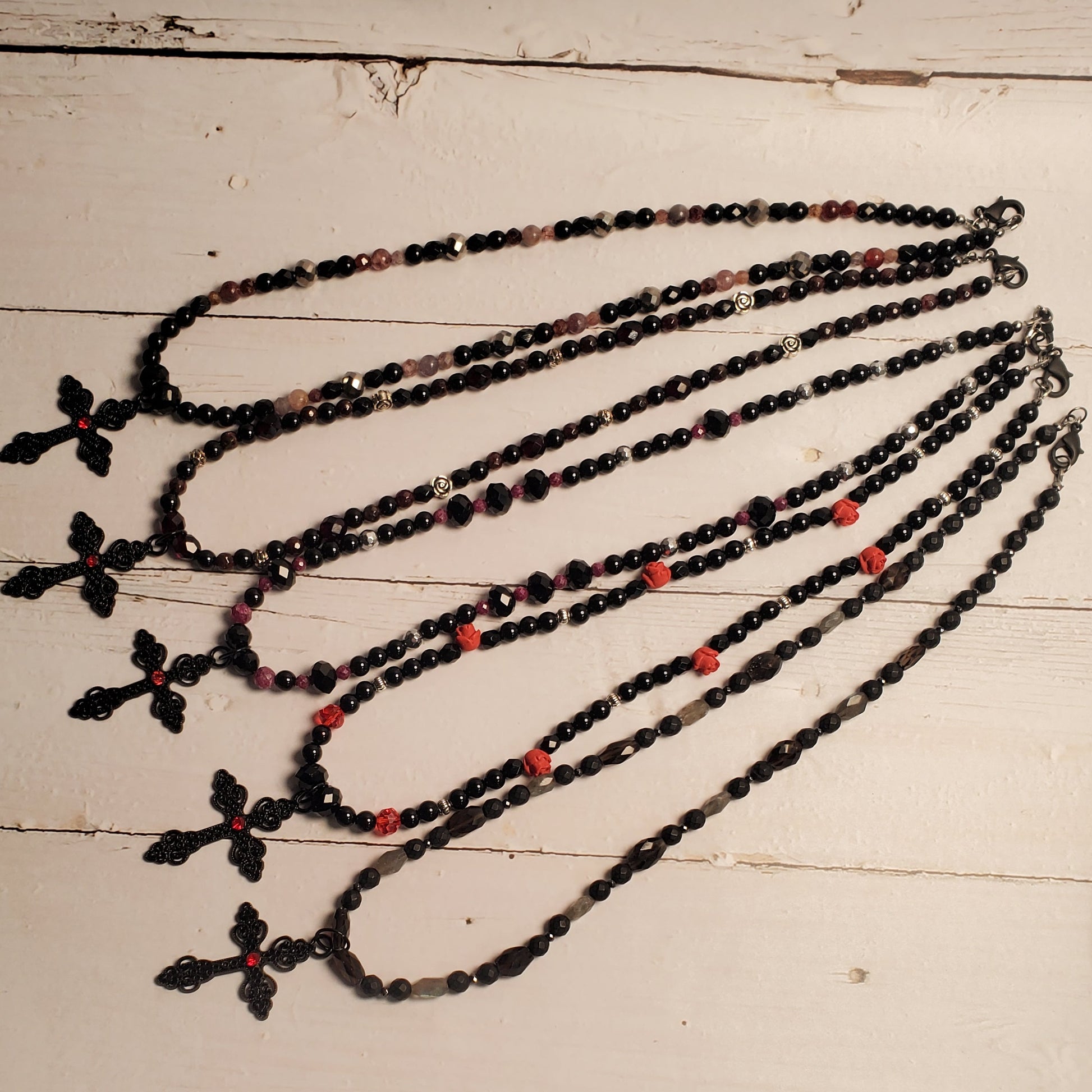 Five beaded necklaces with black and pink beads and cross pendants are laid out on a white wooden table. A person's legs and feet are visible at the bottom of the frame.