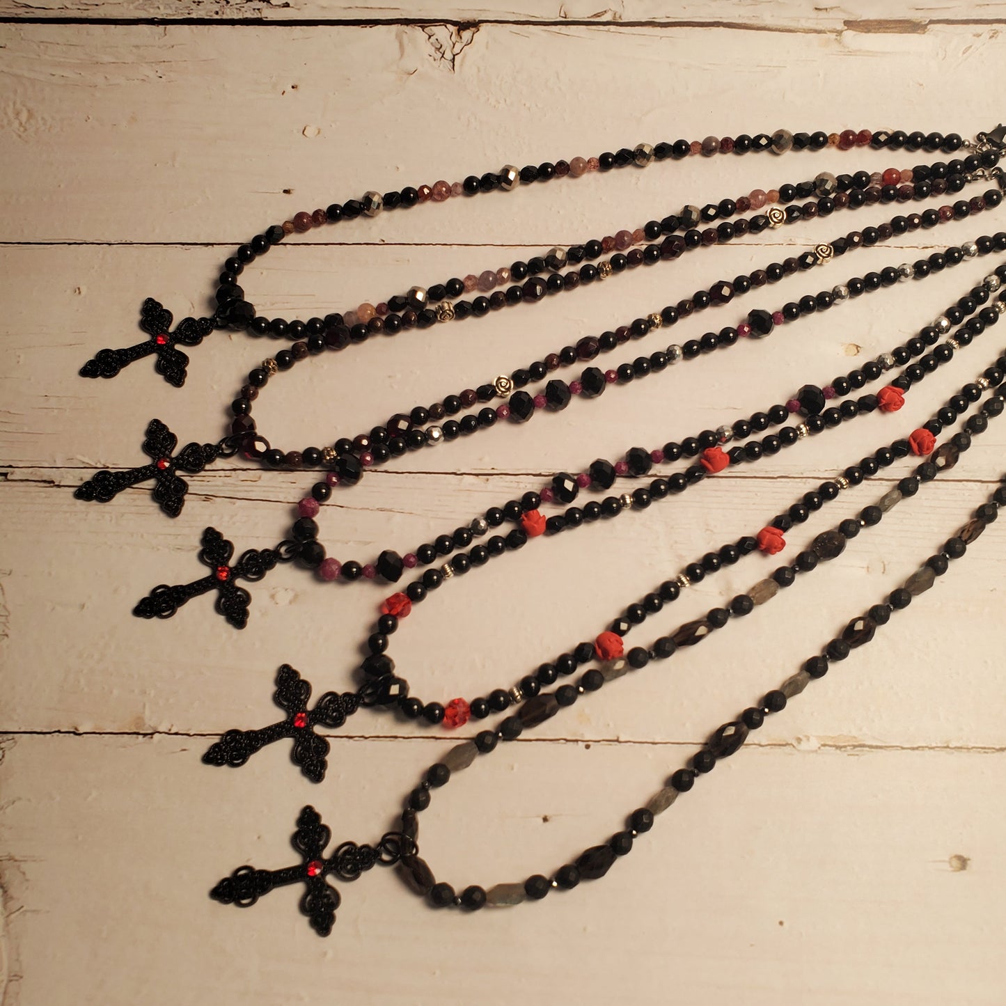 Five long beaded necklaces with black cross pendants are arranged on a distressed white wooden surface. The necklaces feature black faceted beads, small red beads, and some translucent pinkish-red beads.