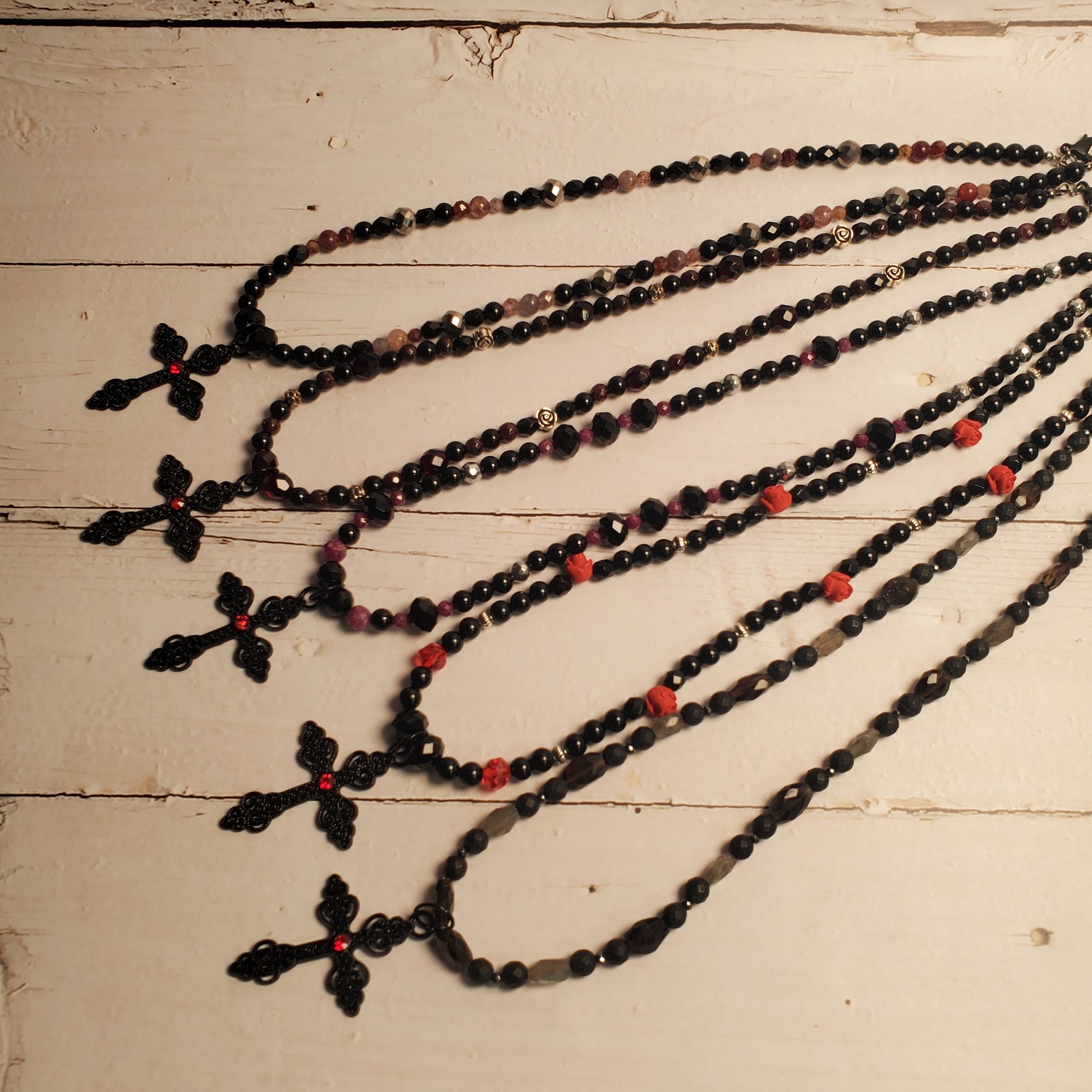 Five long beaded necklaces with black cross pendants are arranged on a distressed white wooden surface. The necklaces feature black faceted beads, small red beads, and some translucent pinkish-red beads.