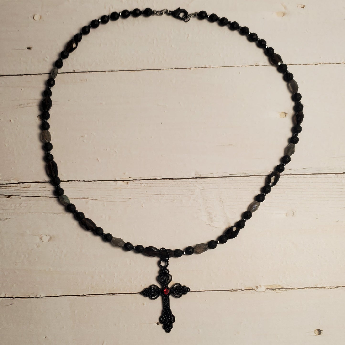 A black beaded necklace with a matching cross pendant, laid flat on a distressed white wooden surface.