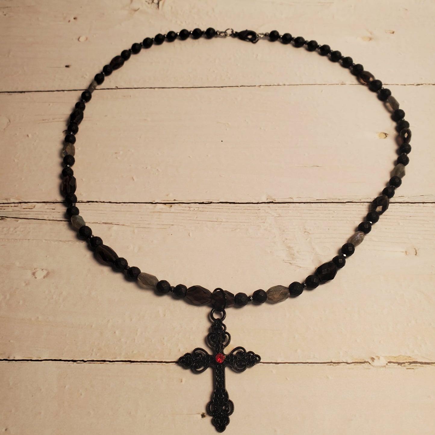 A black beaded necklace with a black cross pendant featuring a small red center detail, displayed on a light beige wooden surface.
