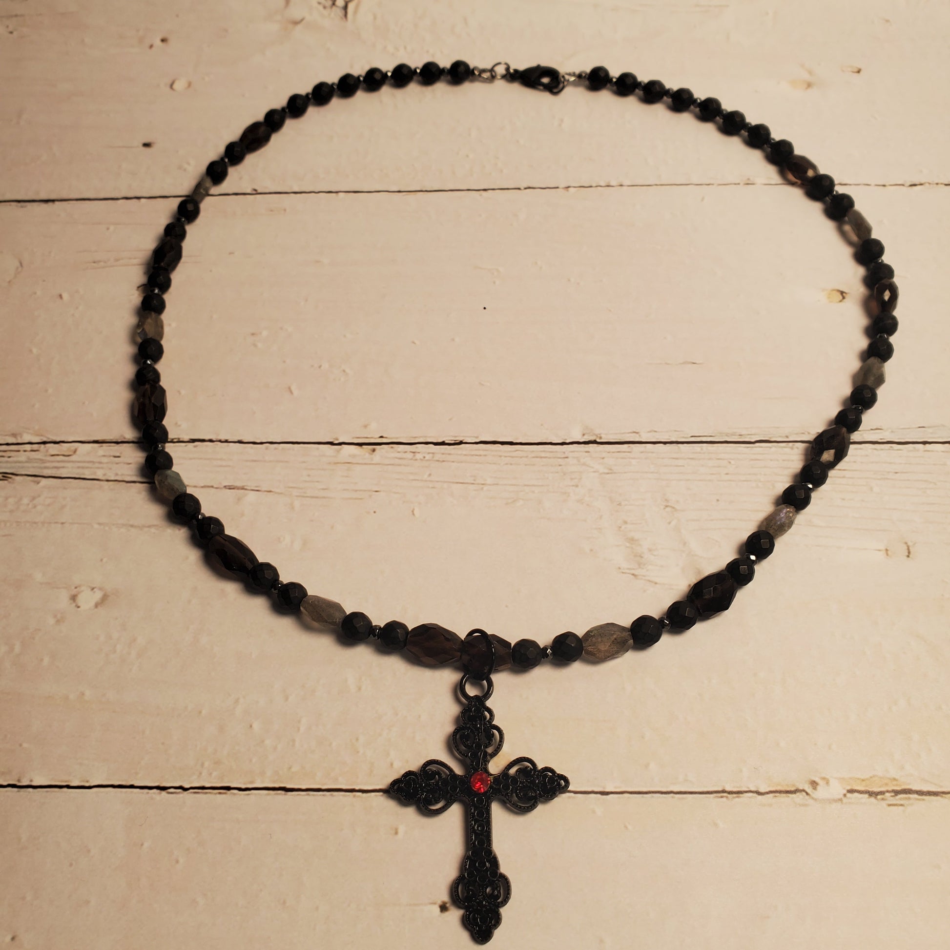 A black beaded necklace with a black cross pendant featuring a small red center detail, displayed on a light beige wooden surface.