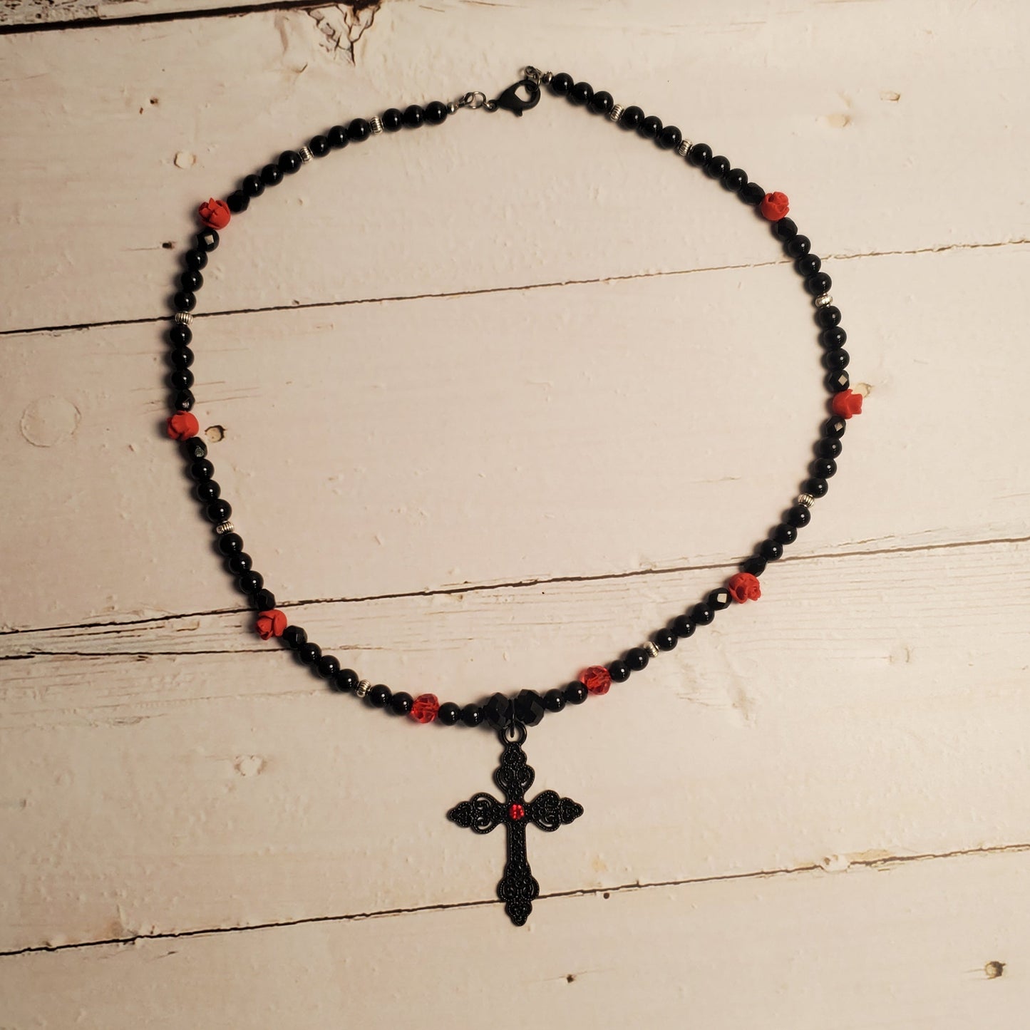 A beaded necklace with black beads, small red accent beads, and a black cross pendant, displayed on a distressed white wooden surface.