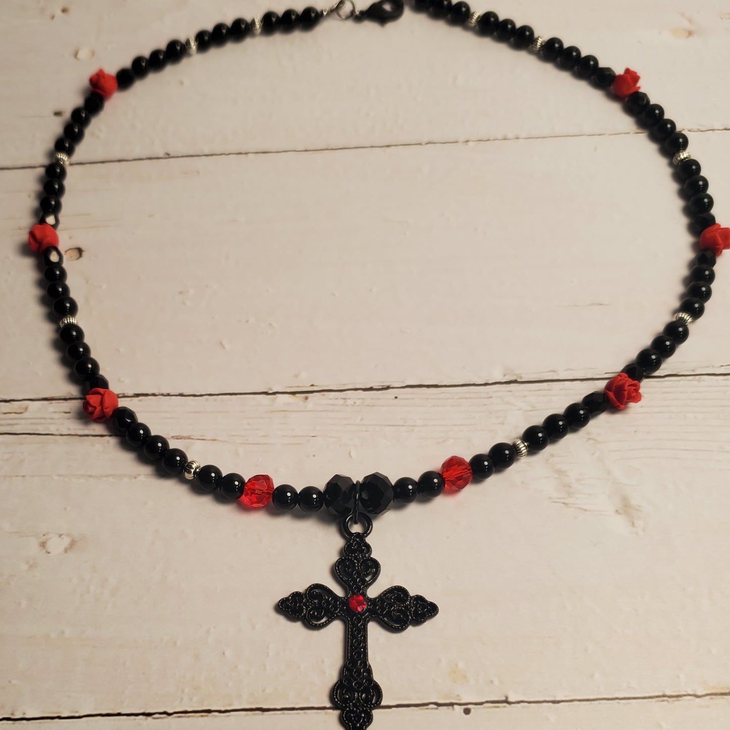 A black beaded necklace with small red accent beads and a black cross pendant, displayed on a light beige wooden surface.
