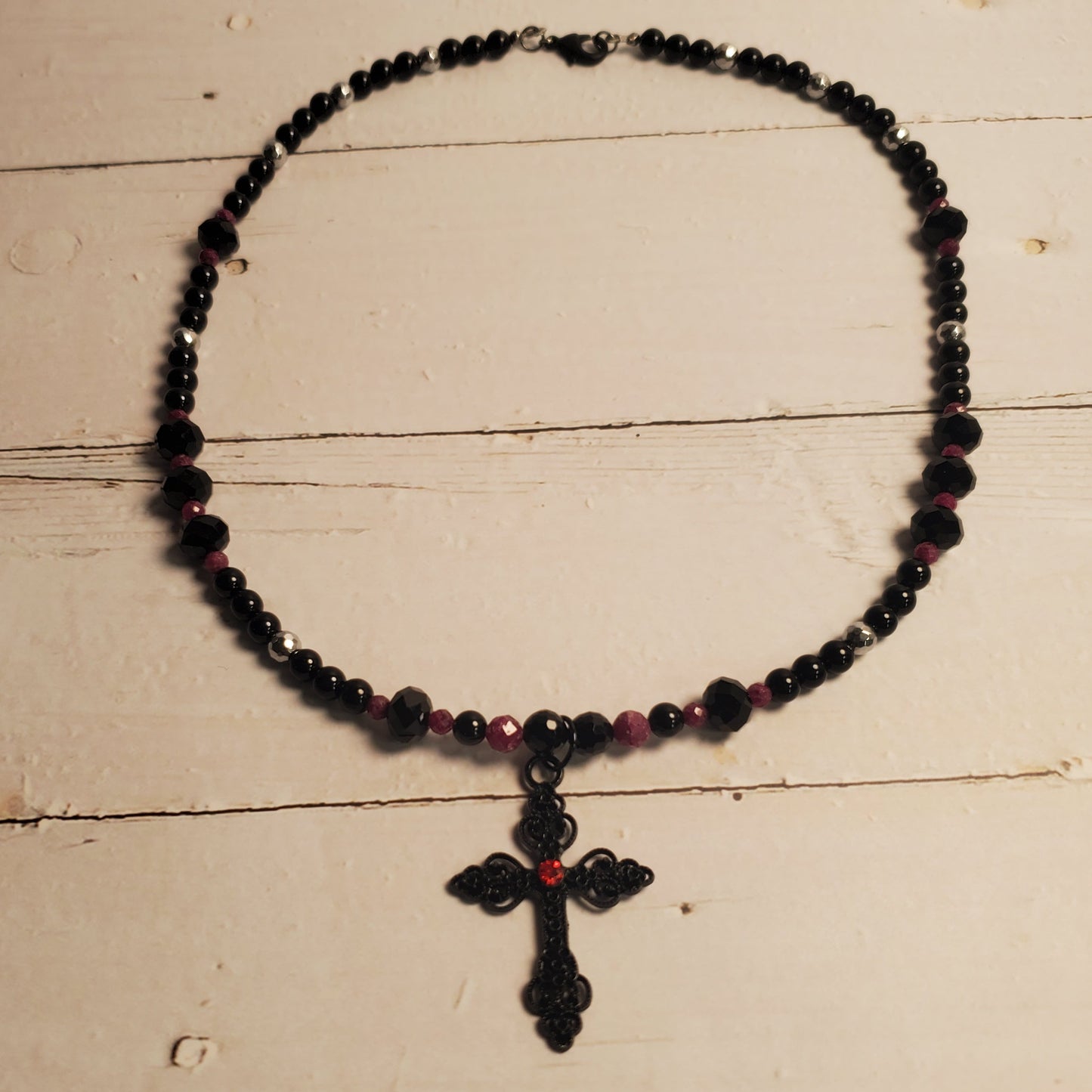 A beaded necklace with a black cross pendant, featuring dark beads and small red accents, displayed on a rustic white wooden surface.