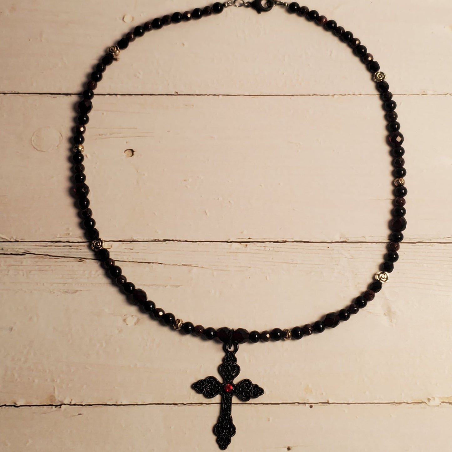 A black beaded necklace with a small cross pendant, laid flat on a distressed white wooden surface.