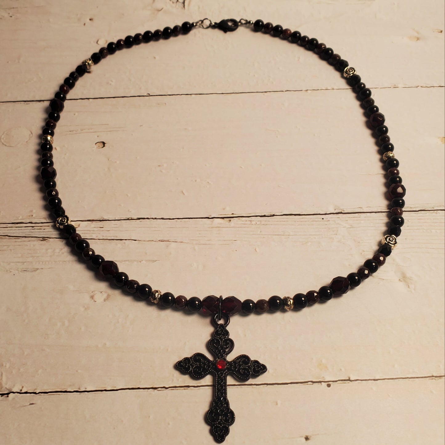 A black beaded necklace with a black cross pendant featuring a small red center detail, displayed on a light beige wooden surface.