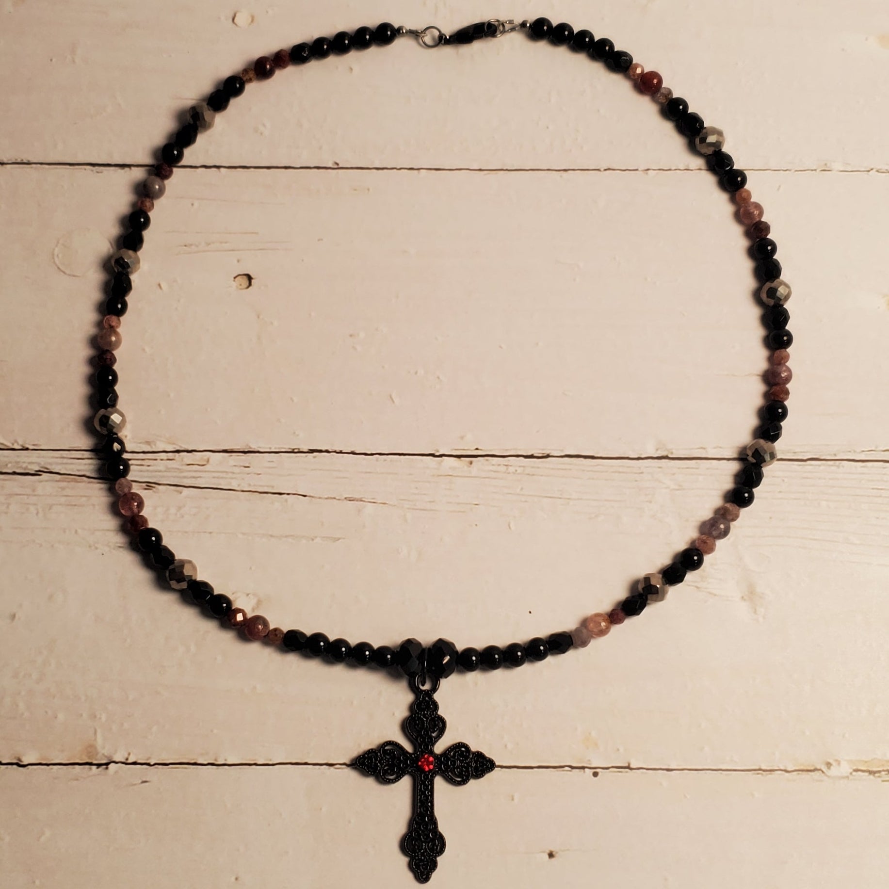 A beaded necklace with a cross pendant, featuring dark beads with subtle pinkish-red accents, displayed against a rustic white wooden background.