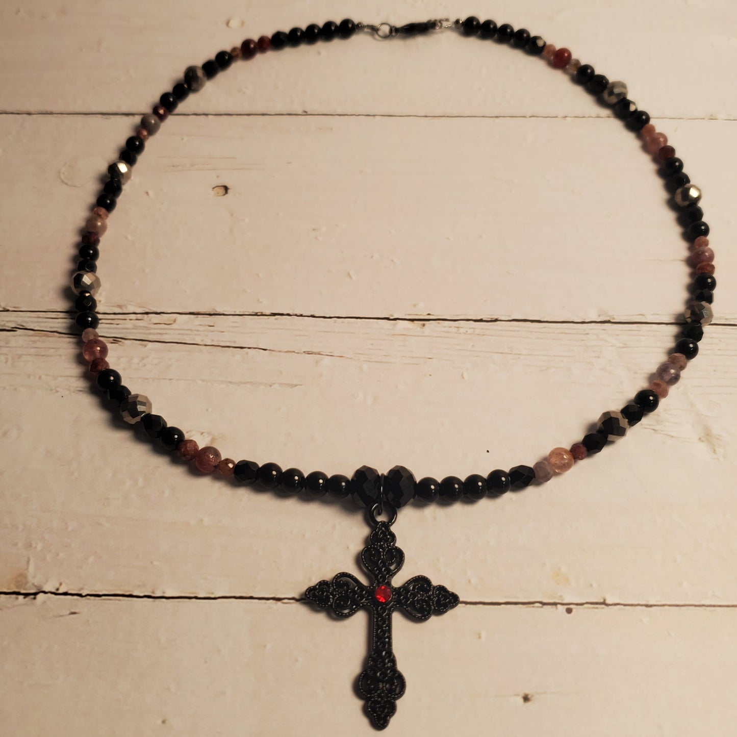 A beaded necklace with a black cross pendant, featuring small dark beads and translucent pinkish-red beads, displayed on a rustic white wooden surface.