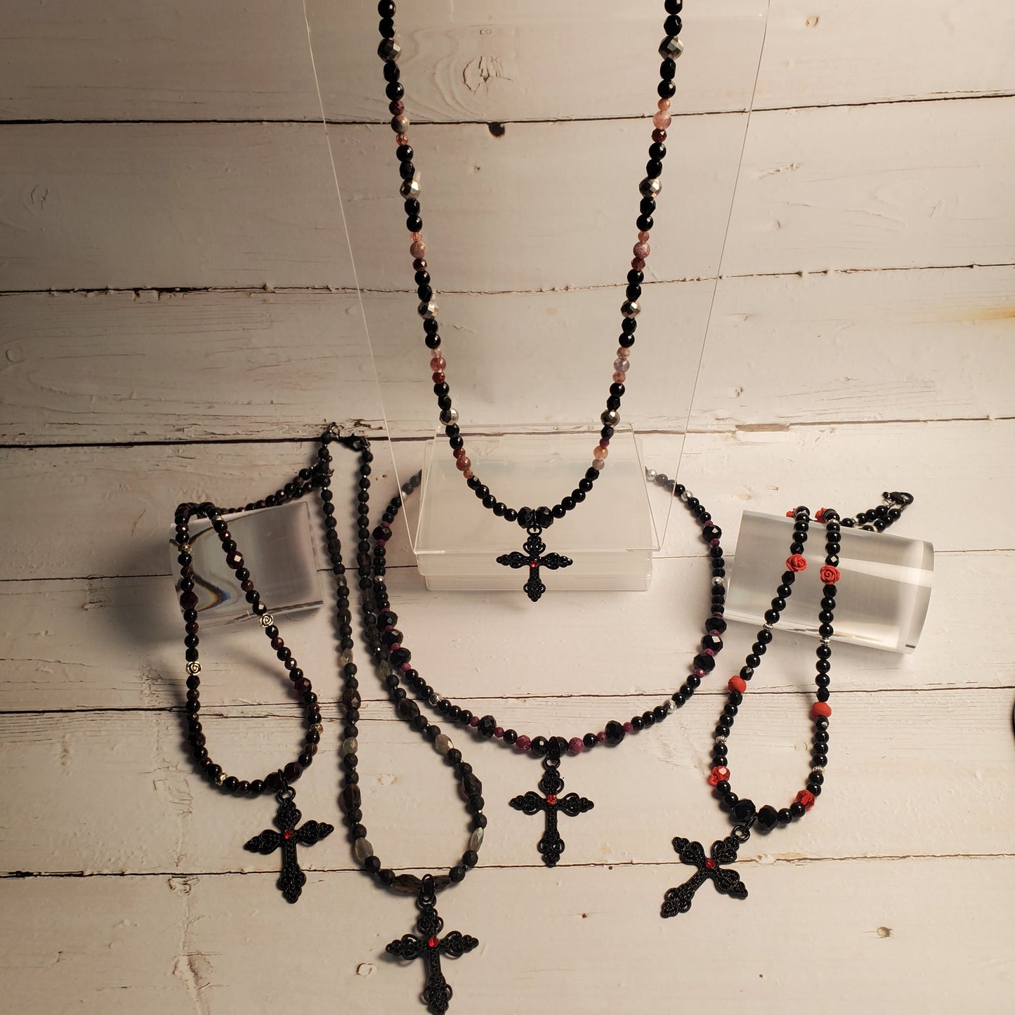 A jewelry set displayed on a white wooden surface, featuring multiple beaded necklaces and bracelets with black beads, small red accents, and cross pendants, shown on clear display stands.