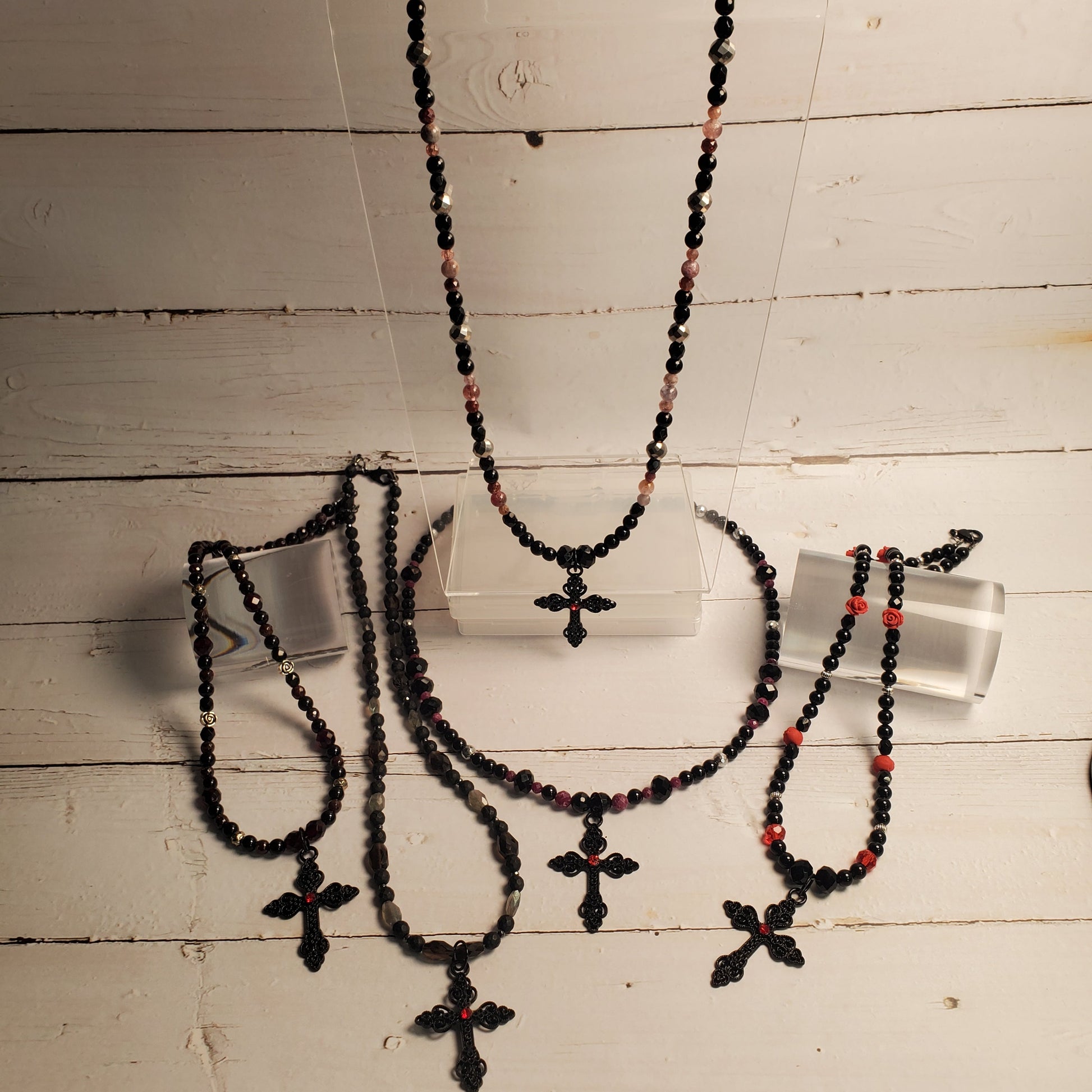 A jewelry set displayed on a white wooden surface, featuring multiple beaded necklaces and bracelets with black beads, small red accents, and cross pendants, shown on clear display stands.