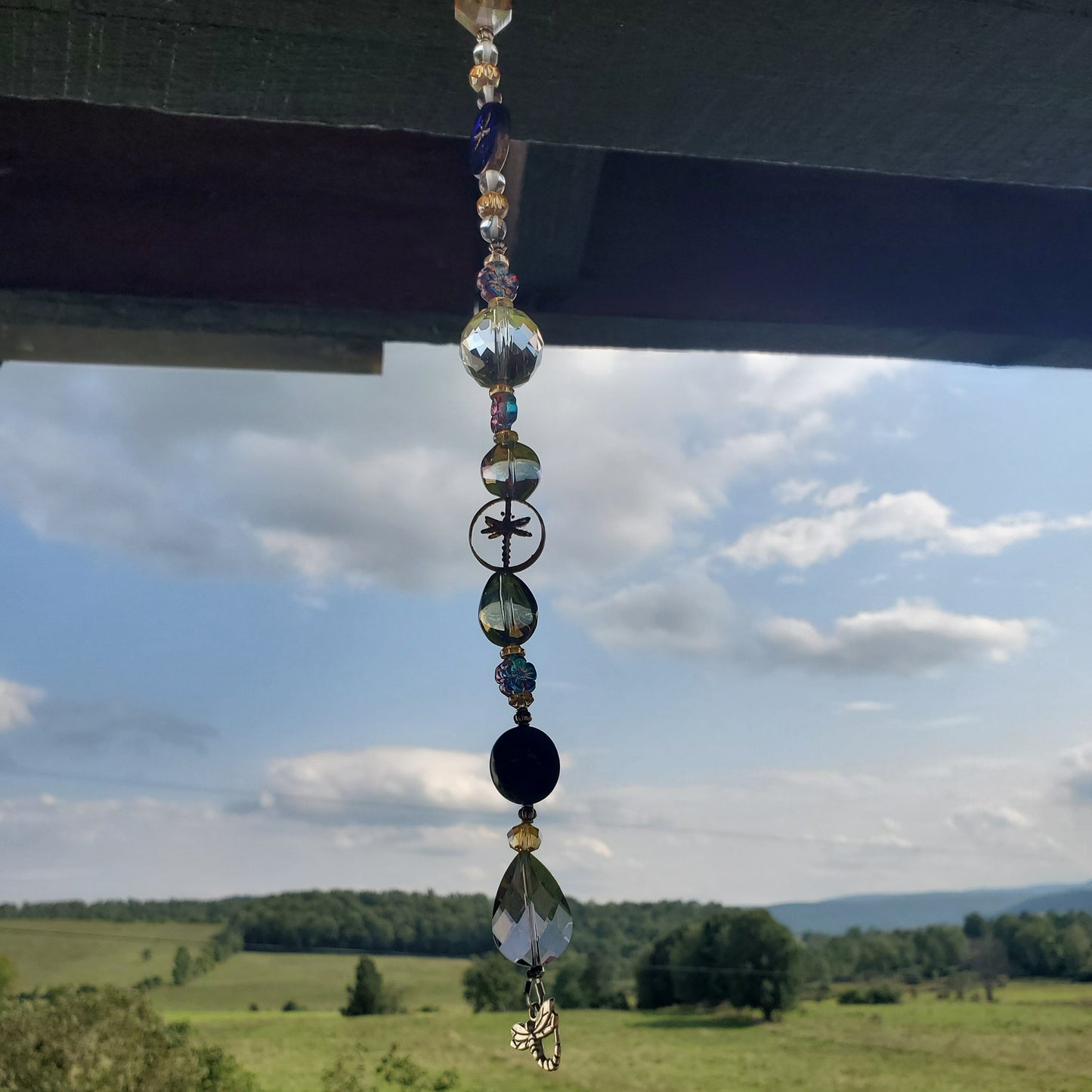 A beaded suncatcherhangs from a dark wooden porch overhang, with a scenic green field, road, and cloudy blue sky in the background.