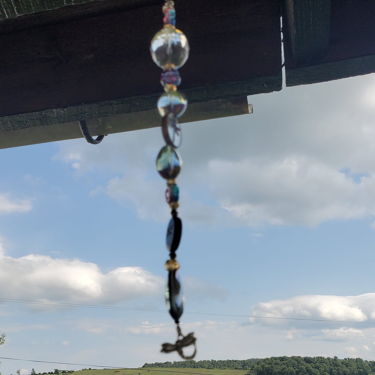 A hanging beaded suncatcher with clear glass and dark beads is suspended from a dark wooden porch overhang, with a green rural landscape and blue sky with clouds in the background.